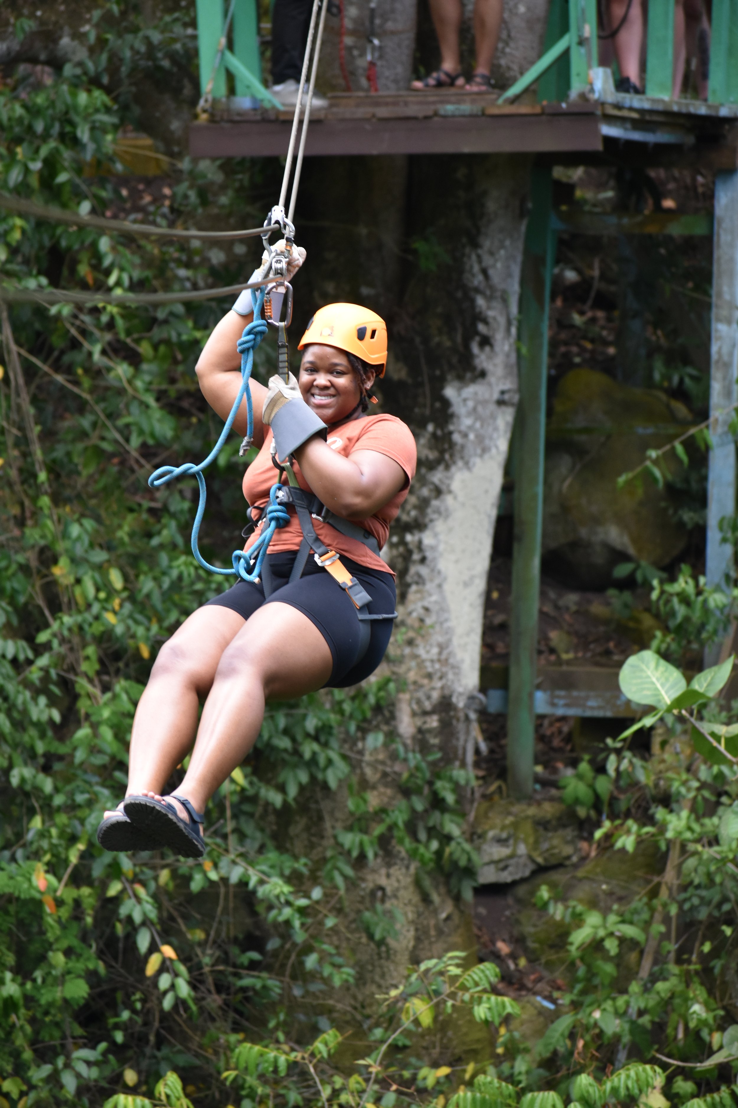 A woman wearing a safety helmet and harness sliding down a zip line through lush green forest.