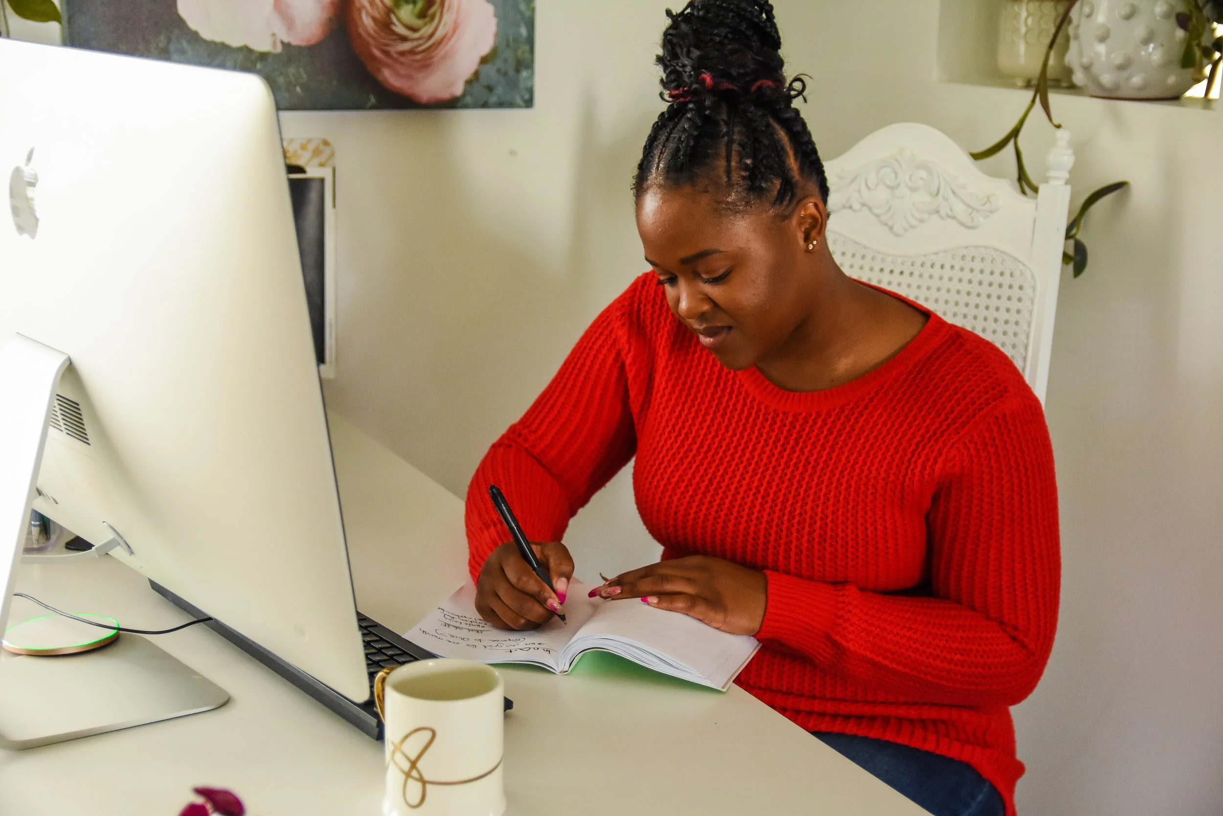 A woman in a red sweater writing in a notebook at her desk next to a computer monitor and a coffee mug.