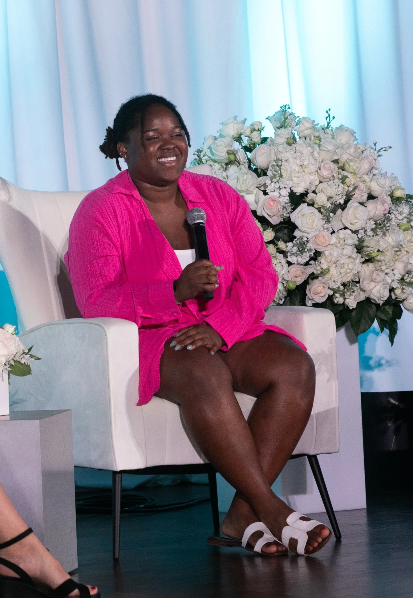 A woman in a pink jacket and white top sitting on a white chair, holding a microphone, with a large bouquet of white flowers behind her.
