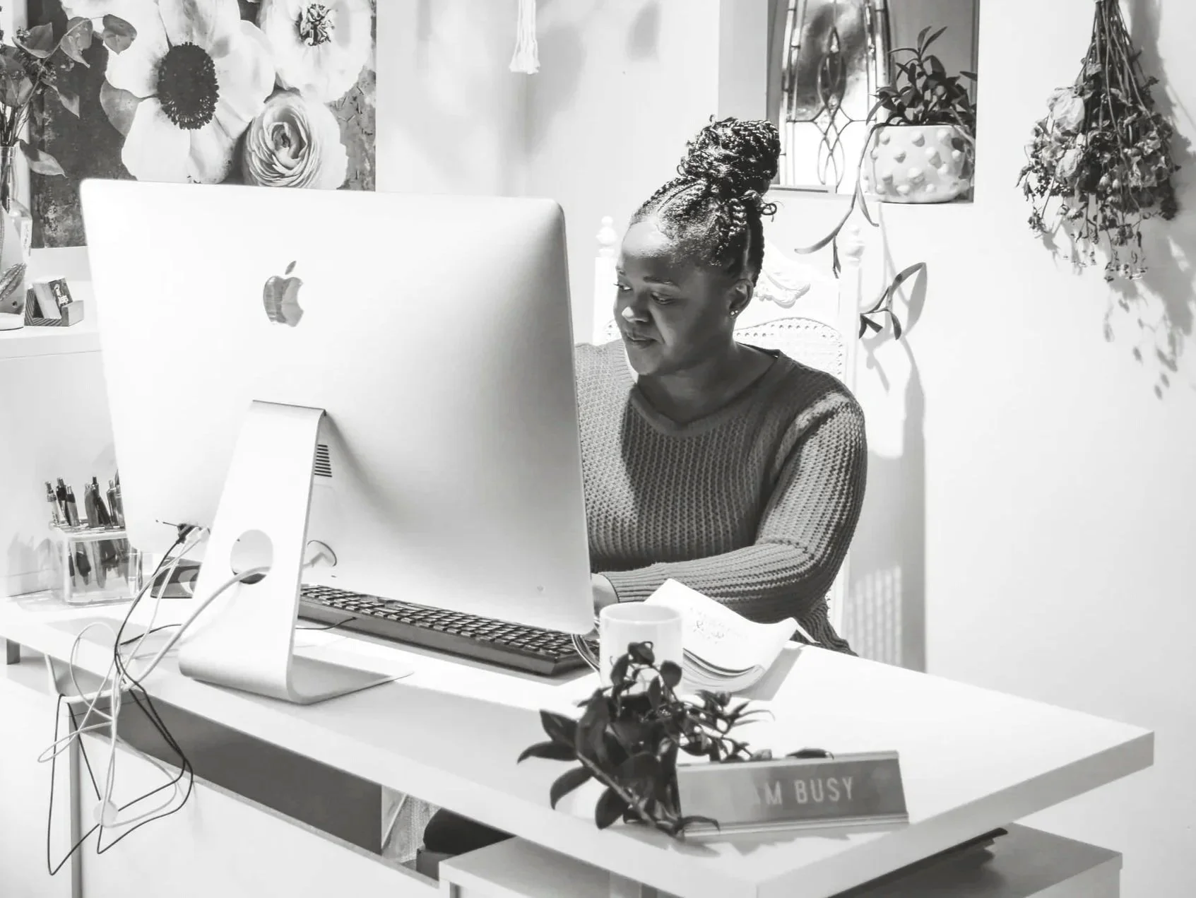 Woman working at a desk with an iMac computer, cup, plant, and a sign that reads 'M BUSY'.
