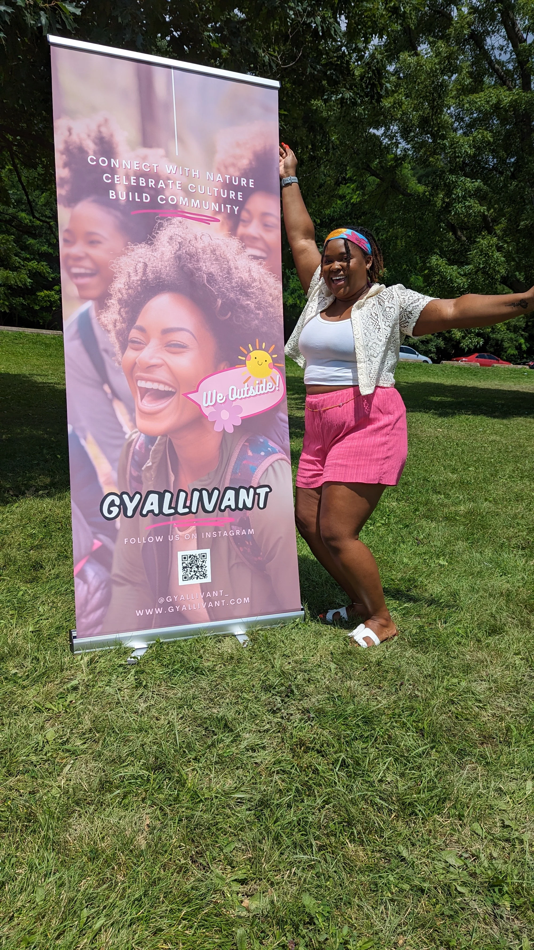 A woman standing outdoors on a grassy area next to a colorful vertical banner. The woman is smiling, wearing a white top, pink shorts, white sandals, and a rainbow-colored headband. The banner features a smiling woman with curly hair, with text promo