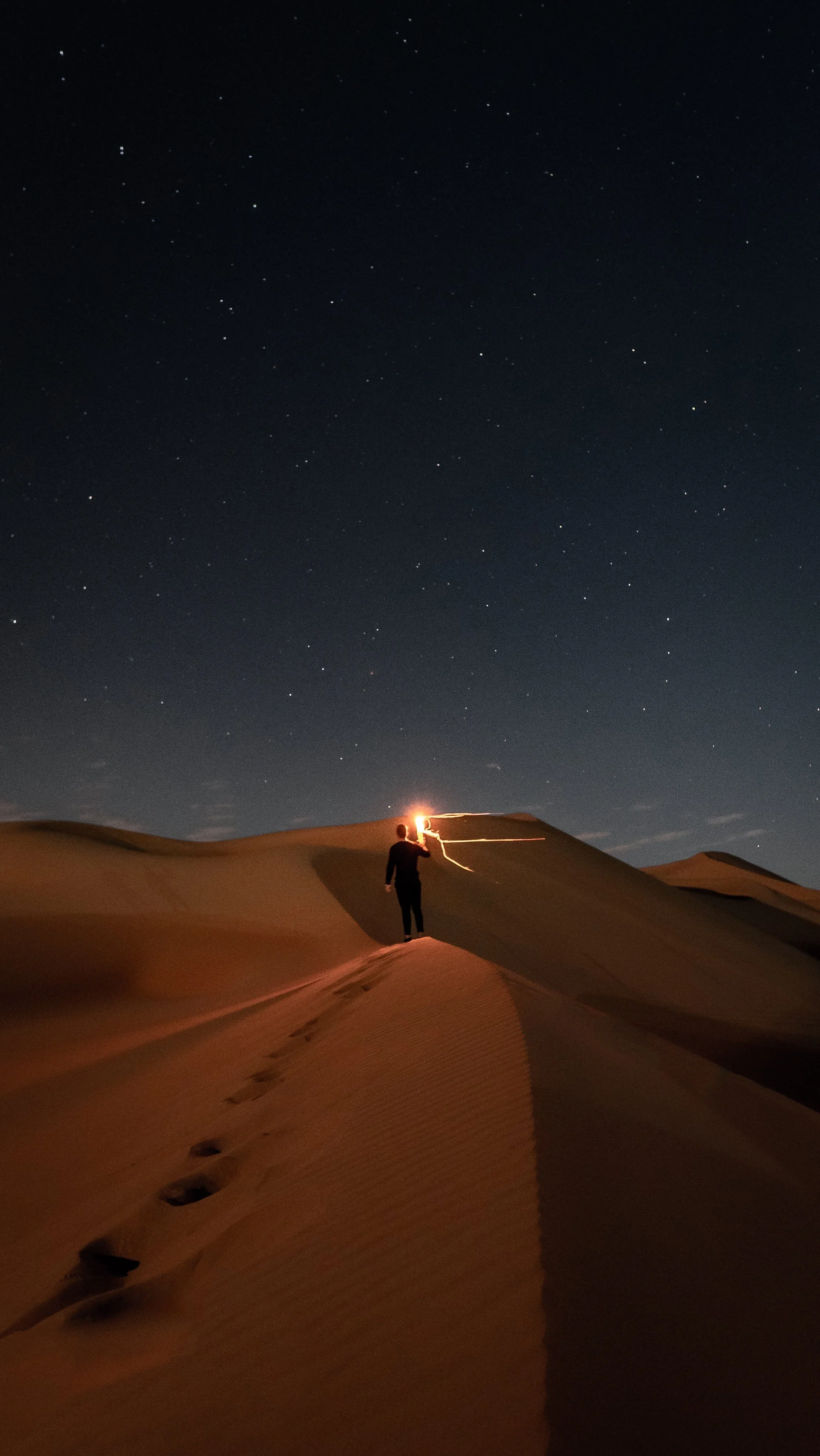 Person walking on sand dunes at night with starry sky overhead, holding a sparkler creating light streaks.