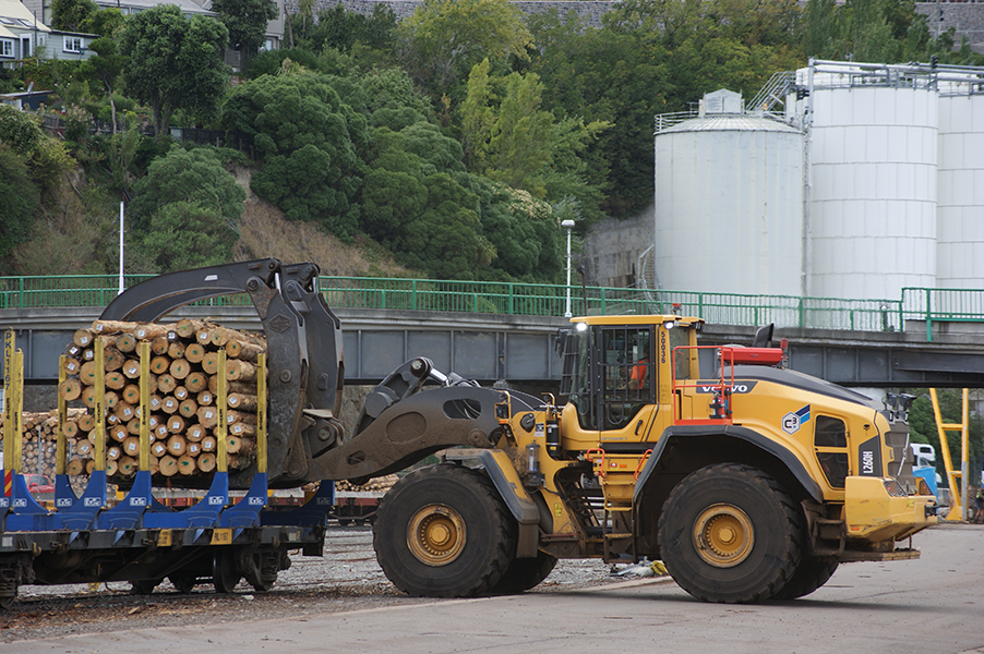 A large yellow loader with black wheels and cabin moving a load of cut logs onto a blue flatbed railcar at a lumber yard, with industrial silos and green foliage in the background.