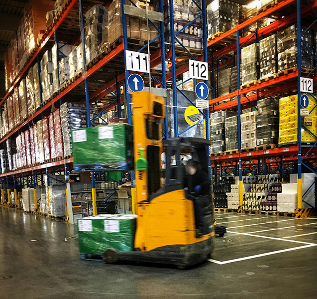 A warehouse with tall shelves stocked with boxes and pallets. A worker operating a yellow forklift is moving green boxes on the warehouse floor.