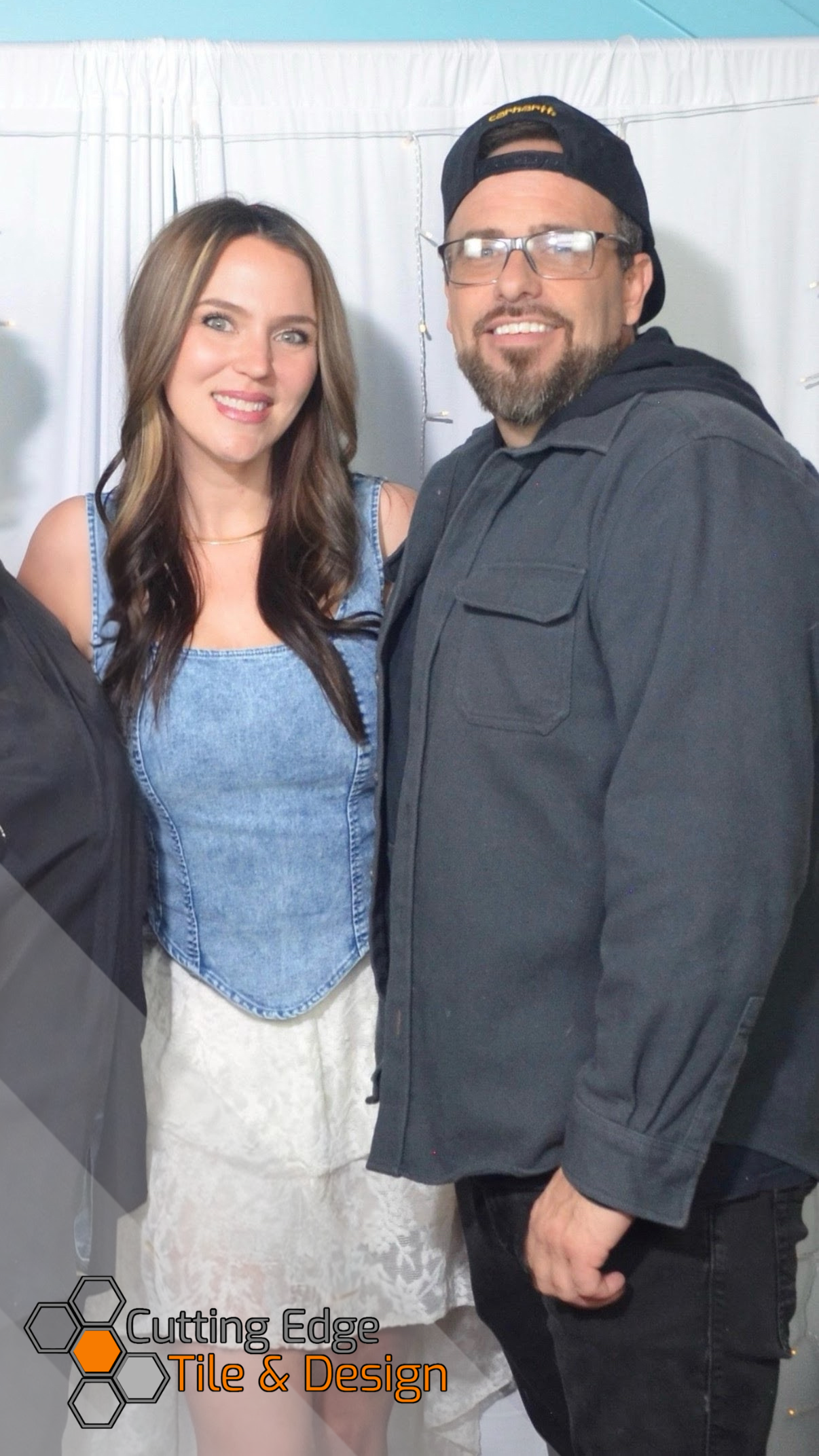 A woman with long brown hair wearing a denim top and a lace skirt standing next to a bearded man with glasses and a backwards black cap, both smiling indoors against a white backdrop with string lights.