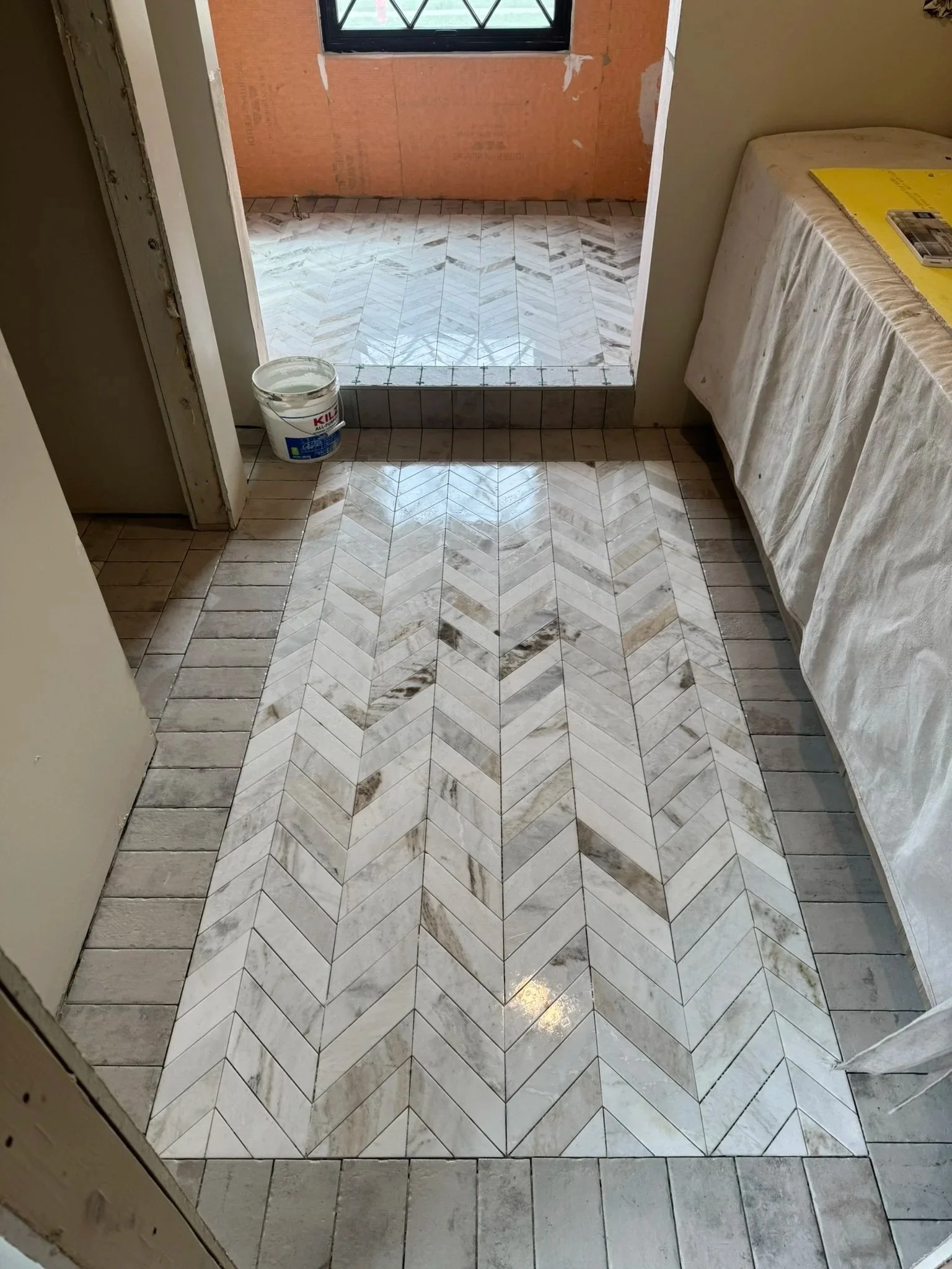 Tile flooring under construction with a chevron pattern in the center, surrounded by plain rectangular tiles, in a room with a window and construction materials.