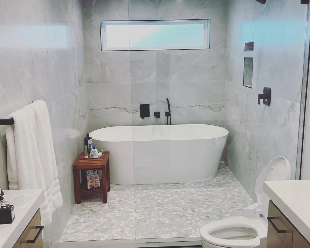 Modern bathroom with marble walls and floor, freestanding bathtub under a window, toilet on the right, small wooden table with toiletries on the left, towel hanging on a rack, and a glass shower door.