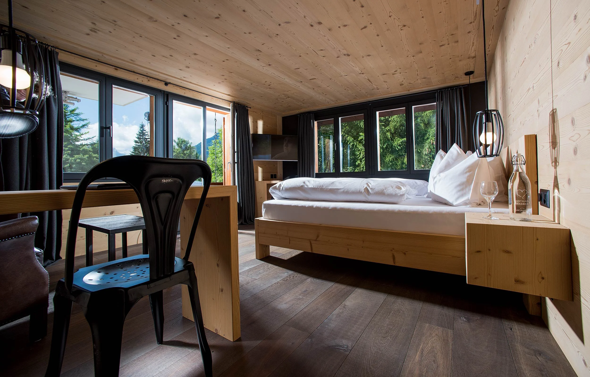 Modern bedroom with large windows, wooden decor, a bed, table with chair, and a lamp.