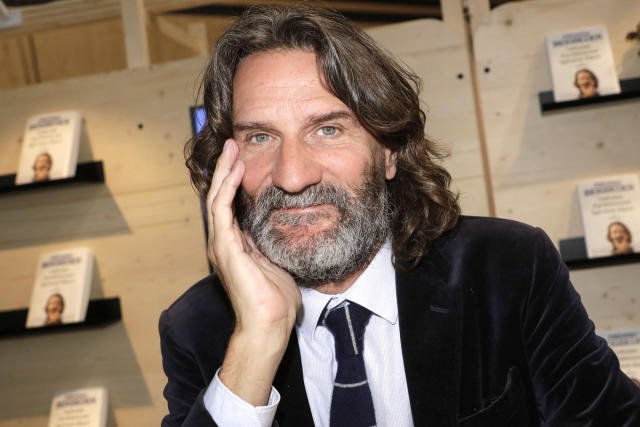 A man with a beard and long hair smiling, resting his head on his hand, books on shelves in the background.