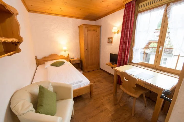 Cozy hotel room with single bed, wooden furniture, small TV, armchair, desk, and window with red curtains.