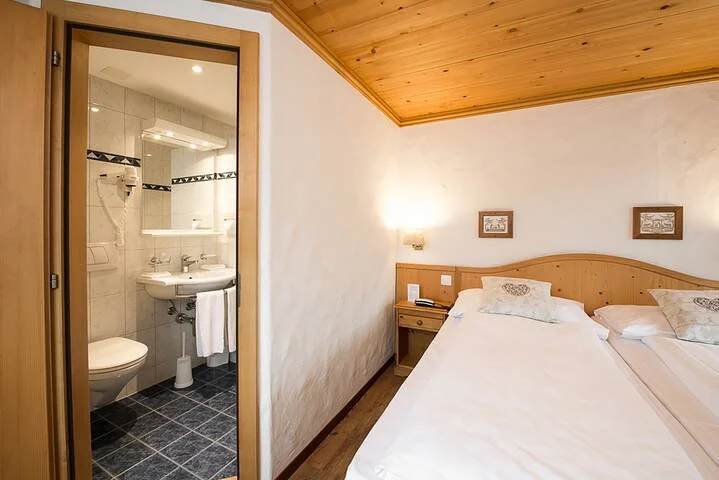 Hotel room with twin beds and en-suite bathroom featuring a tiled shower, sink, and toilet; wooden ceiling and walls, nightstand with lamp and phone.
