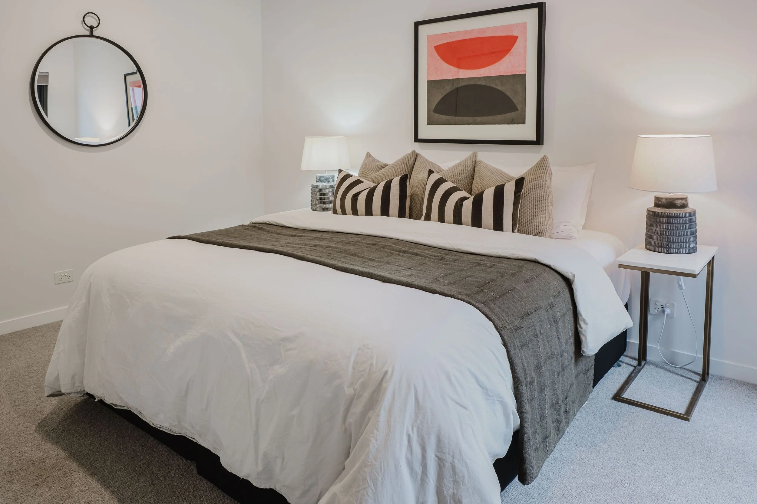 A modern bedroom with a neatly made bed, dark gray blanket, and decorative pillows. There are two white nightstands with matching lamps, a framed abstract artwork above the bed, and a round mirror on the wall.