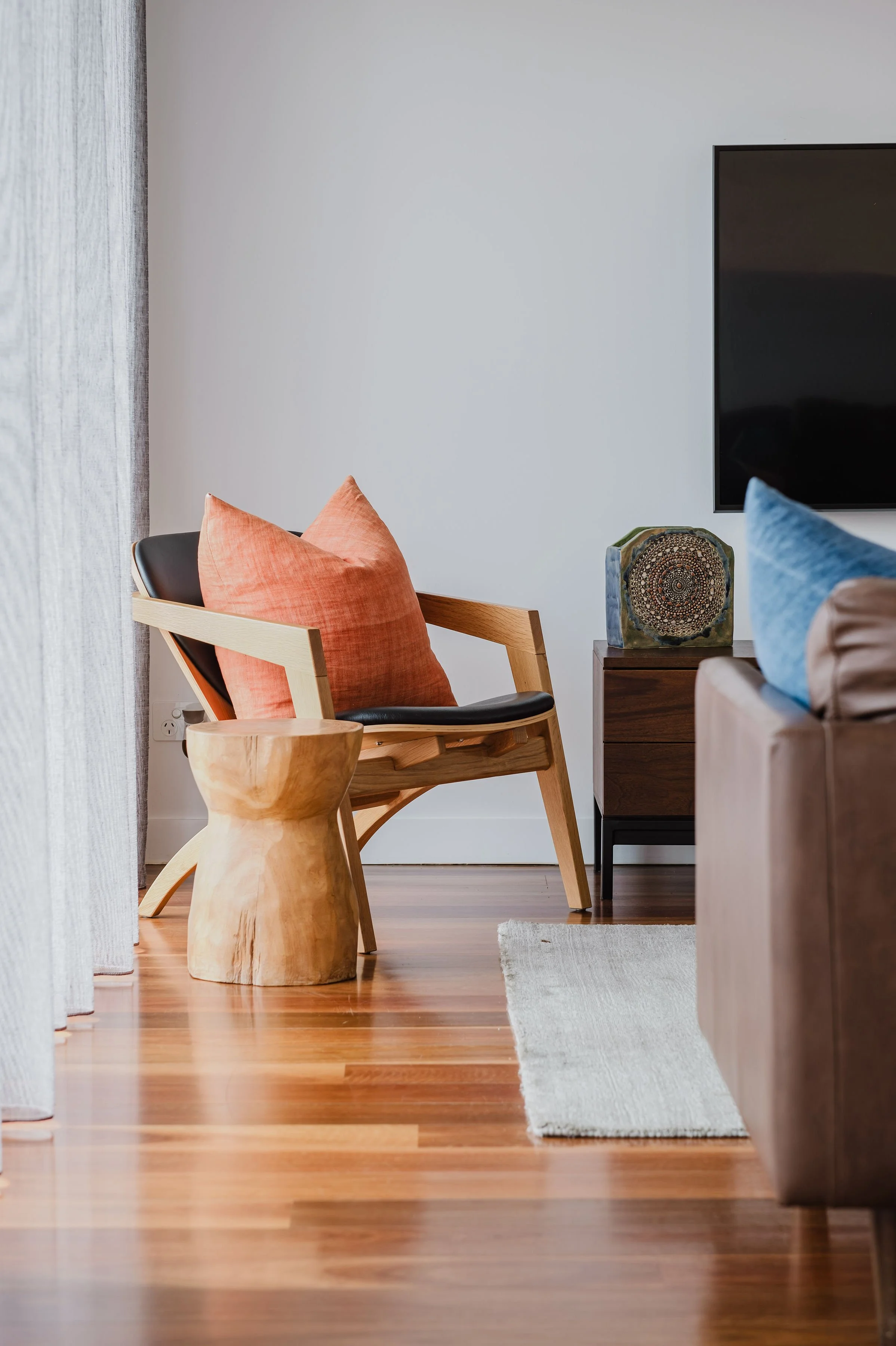A cozy living room corner with a wooden chair featuring black cushions and an orange pillow, a wooden side table with a decorative stone art piece, a brown sofa with a blue pillow, a beige rug on a polished wooden floor, and a large flat-screen TV mo