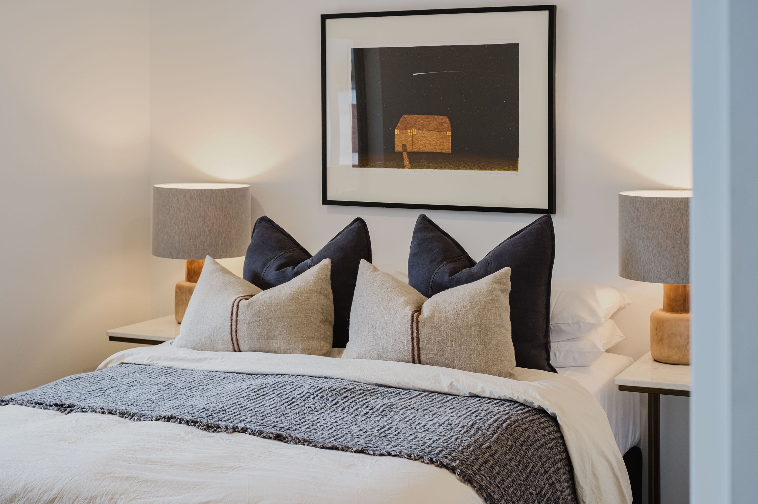 A neatly made bed with a row of four beige pillows and two larger black pillows behind them. The bed has a textured gray blanket folded across the foot. There are two nightstands with wooden lamps on each side of the bed. Above the bed hangs a framed
