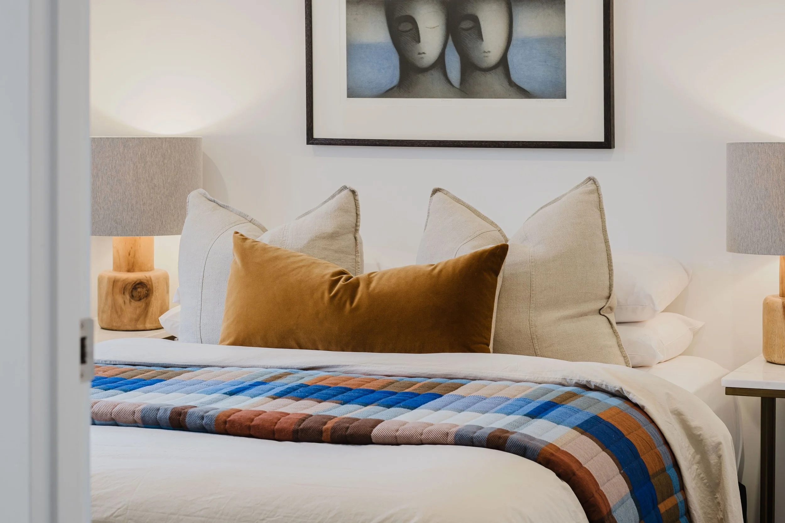 A neatly made bed with beige, white, and brown pillows, a colorful checkered throw blanket, and framed artwork of three faces hangs above it in a cozy bedroom with matching nightstands and lamps.