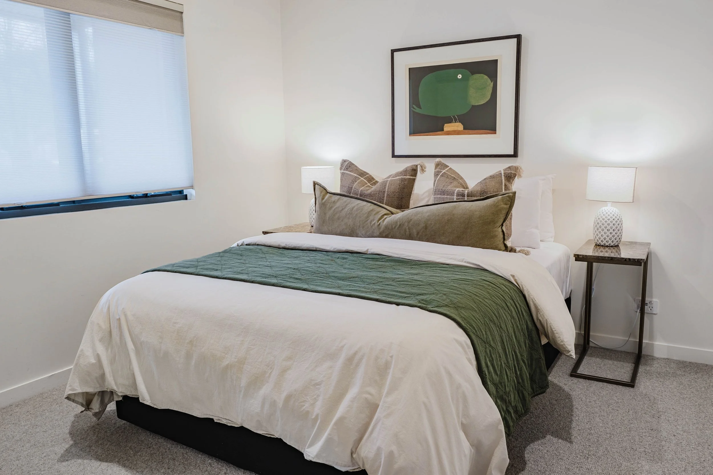 A neatly made bed with a white bedspread and green runner, topped with multiple pillows, in a bedroom with white walls, a window with blinds, and a framed abstract art piece of a green whale above the bed. There are two bedside tables with white lamp
