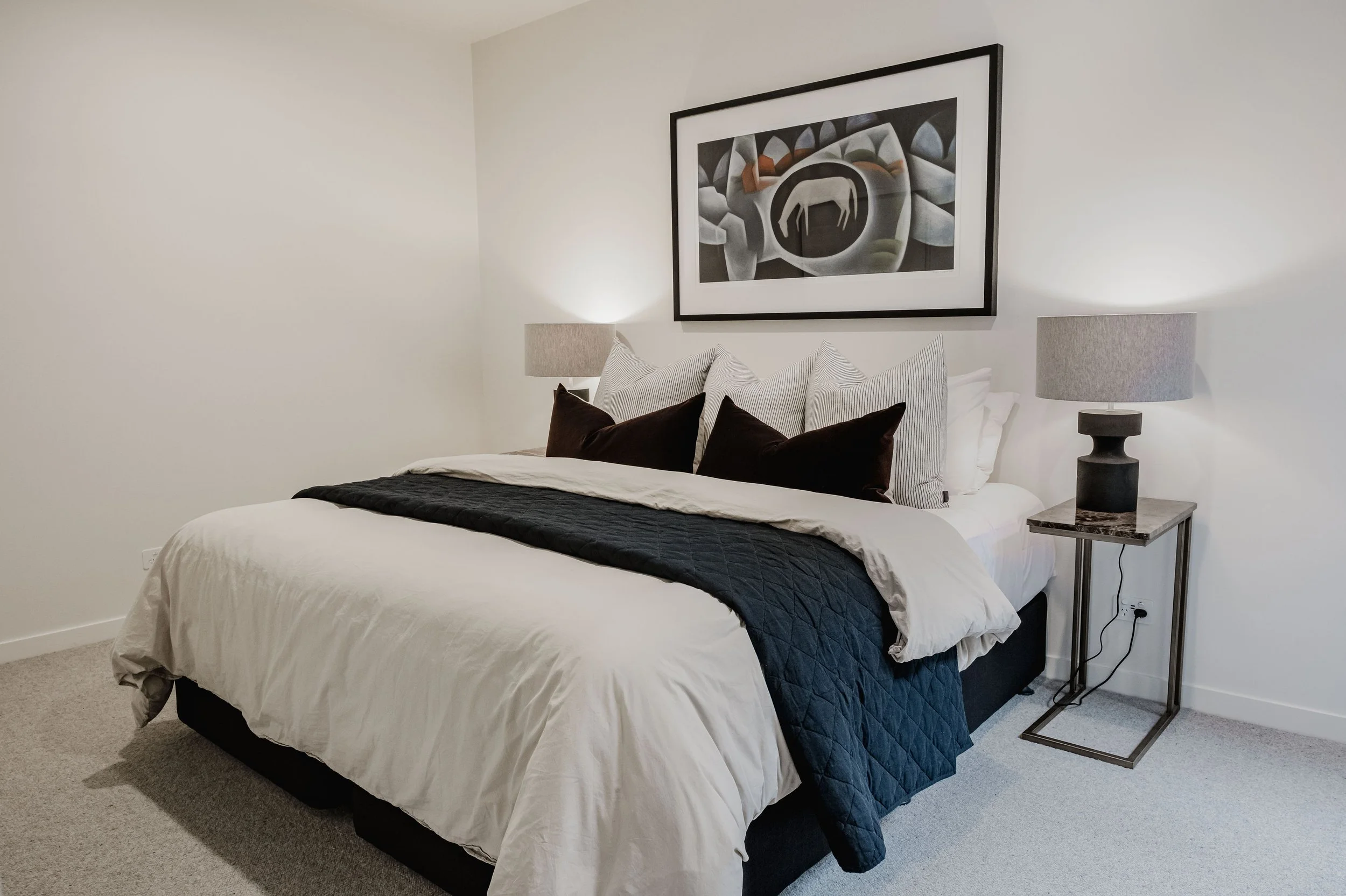 A neatly made bedroom with a white bed covered with white and black blankets and pillows, two grey bedside lamps, and a large framed abstract art piece with geometric shapes hanging above the bed.