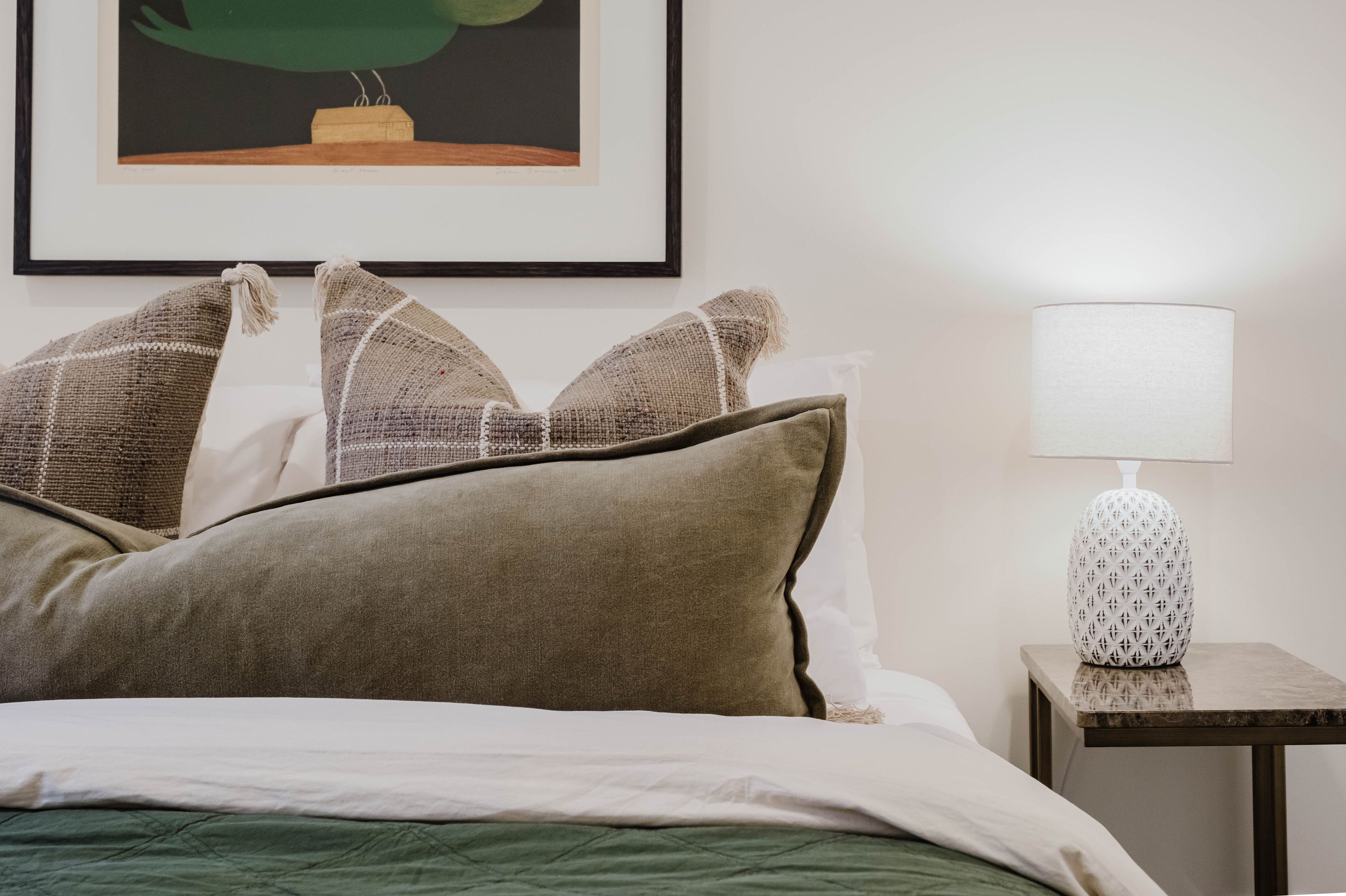 A bedroom with a bed, decorative pillows, a framed artwork, a table lamp, and a nightstand.
