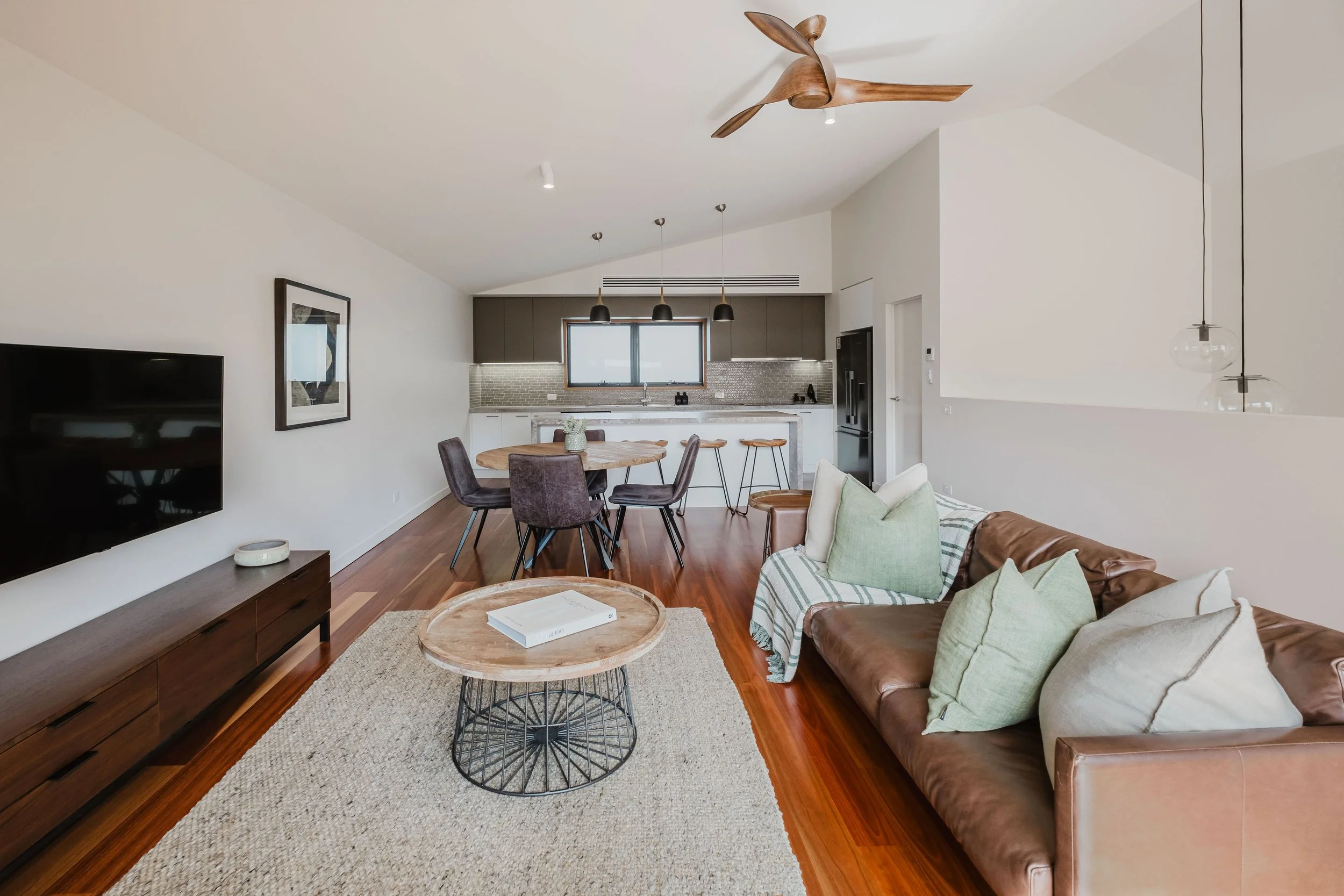 Open-concept living room and kitchen with hardwood floors, a leather sofa with pastel pillows, a wooden coffee table, a television on a wooden stand, a dining table with six chairs, and a modern kitchen with pendant lights, window, and bar stools.