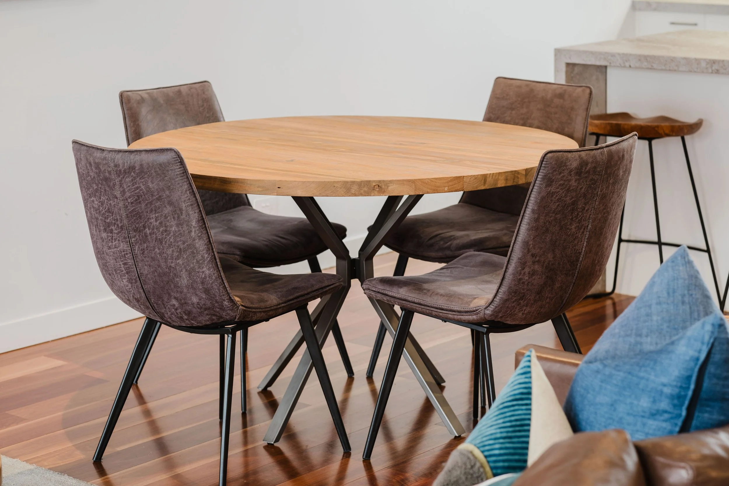 Wooden round dining table with four brown upholstered chairs on a hardwood floor in a room with white walls.