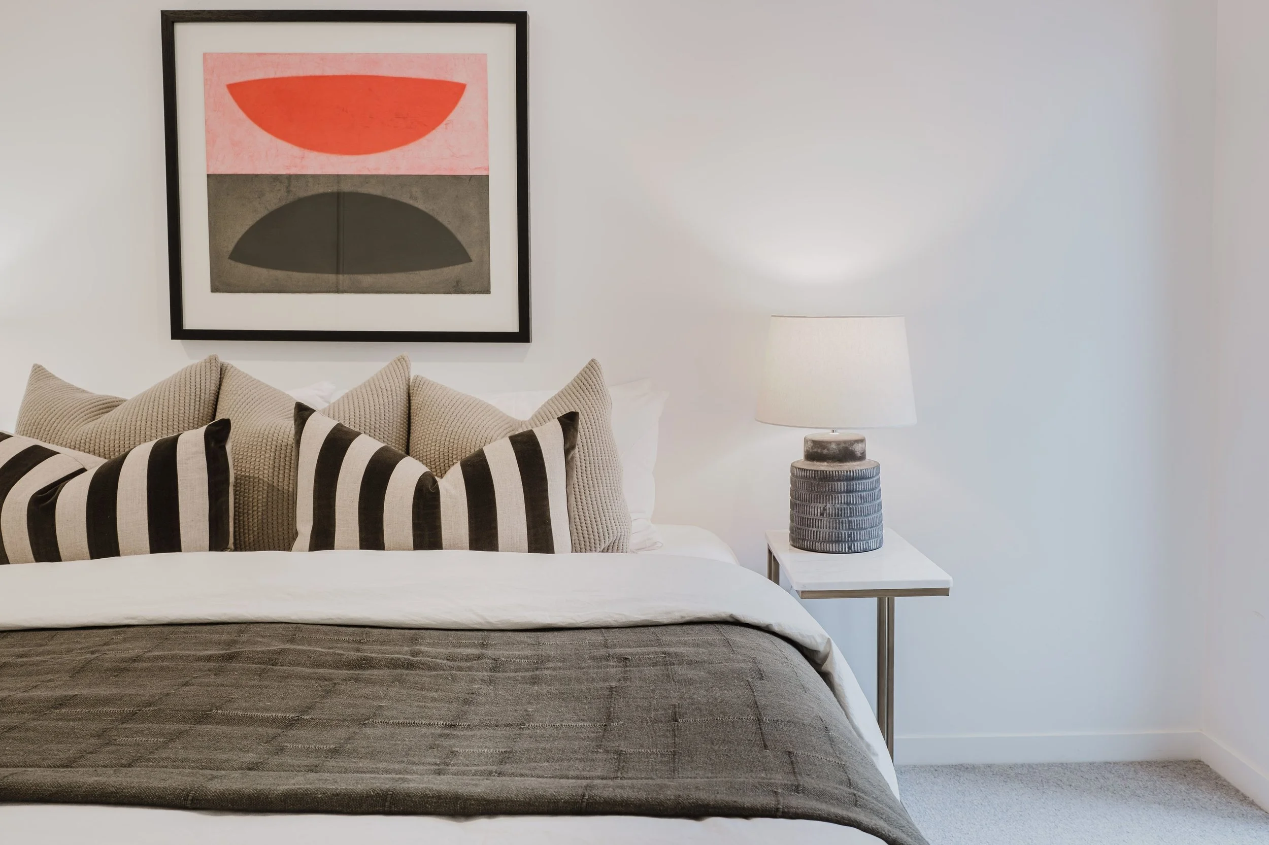 A neatly made bed with multiple beige and black striped pillows, a gray bedspread, and a white blanket. A column-style table lamp with a white shade and a textured gray base is placed on a small white nightstand to the right. A framed abstract art pi