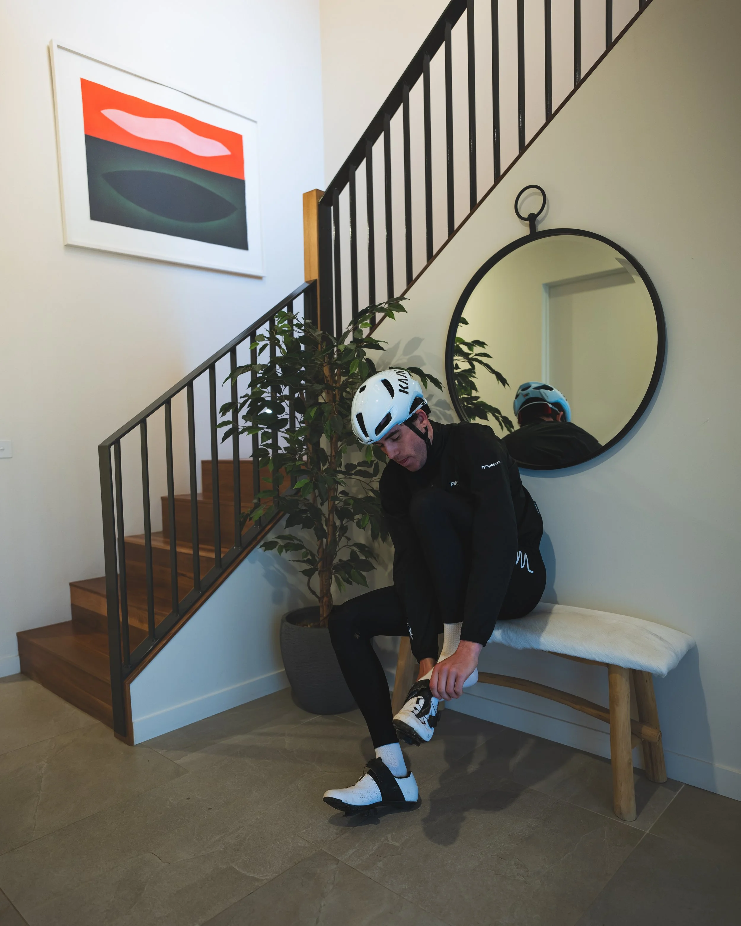 A man wearing a bike helmet sits on a bench by a staircase, putting on cycling shoes, with a round mirror reflecting his back and a large potted plant nearby. An abstract artwork hangs on the wall.