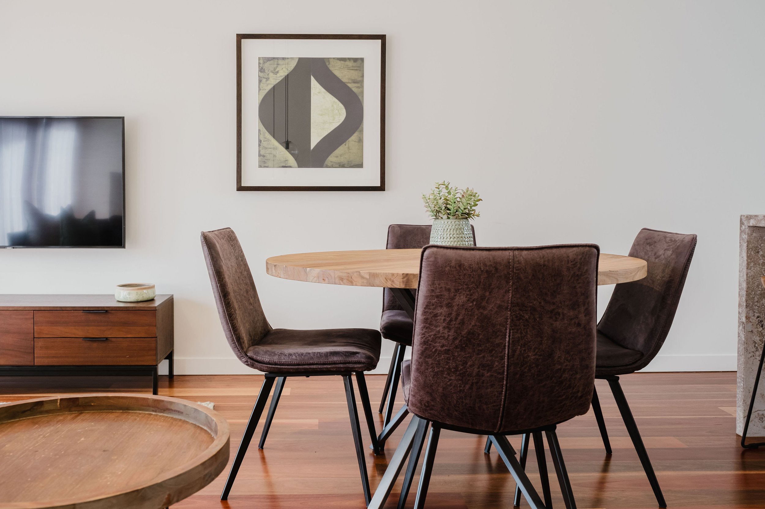 A dining room with a round wooden table surrounded by four brown upholstered chairs, a small plant in a decorative pot on the table, a wooden TV stand with a small dish, a wall-mounted flat-screen TV, and a framed abstract artwork on a white wall.