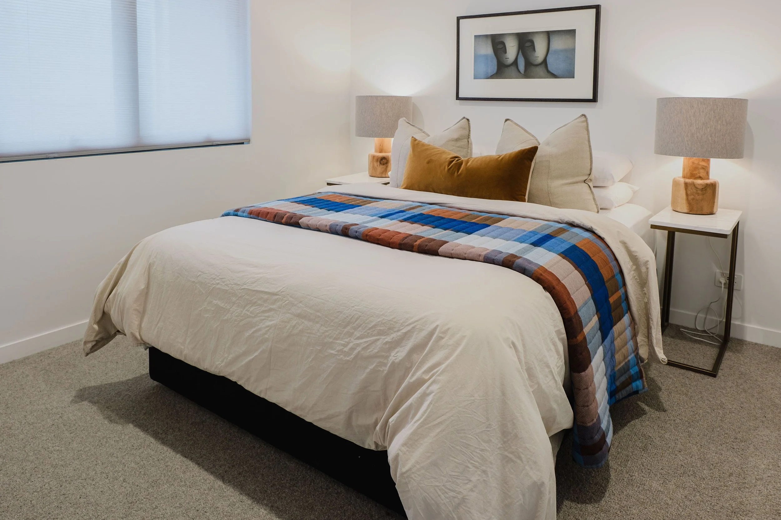 A bedroom with a neatly made bed, beige bedspread, colorful plaid blanket, and several pillows, including a gold one. There are two nightstands with lamps and a framed artwork featuring two faces above the bed. A window with closed blinds is on the l