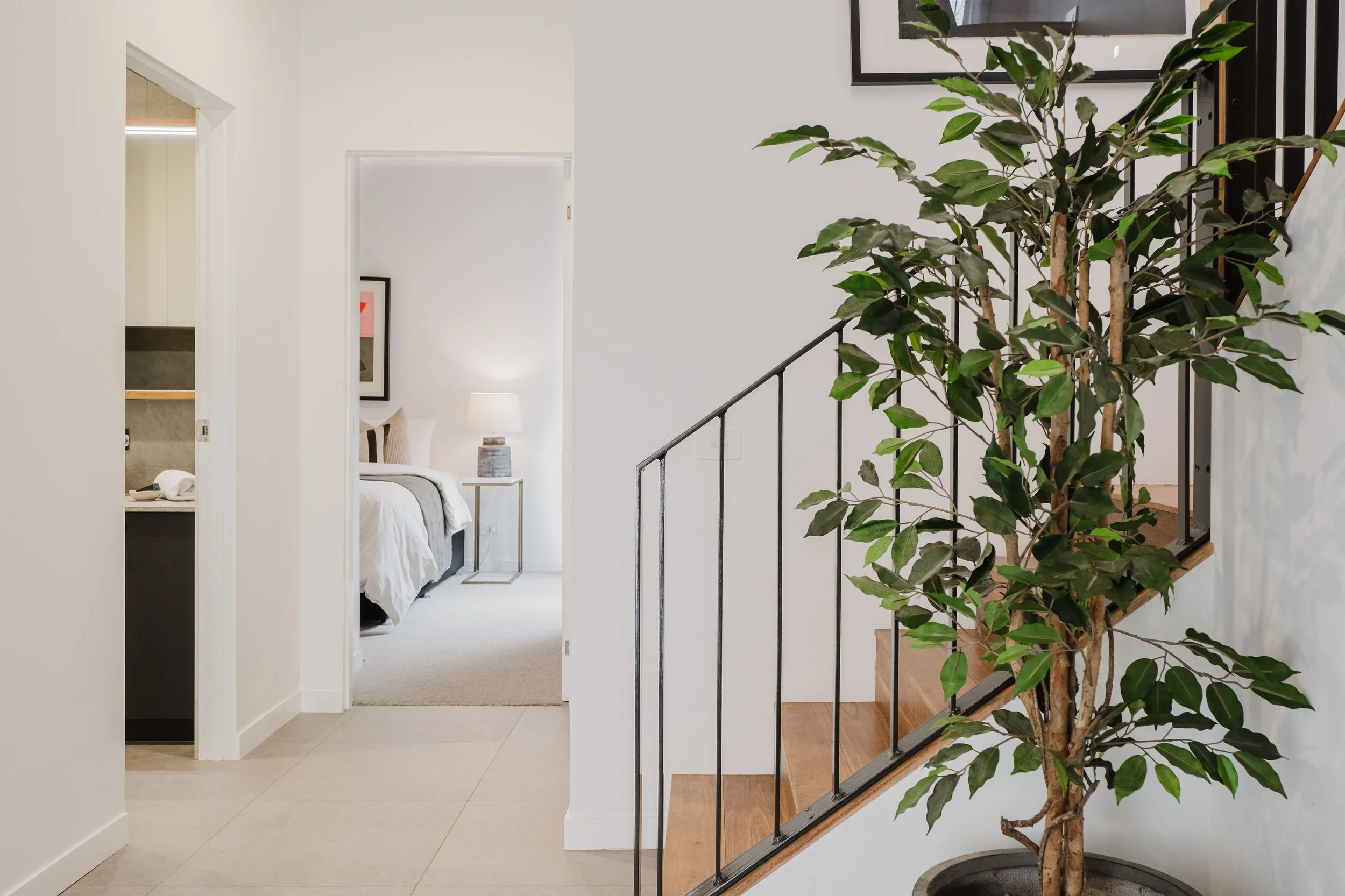 Interior view of a modern home, showing a staircase with black railing, a large indoor potted tree, and a glimpse into a bedroom with a bed, nightstand, lamp, and a framed picture on the wall.
