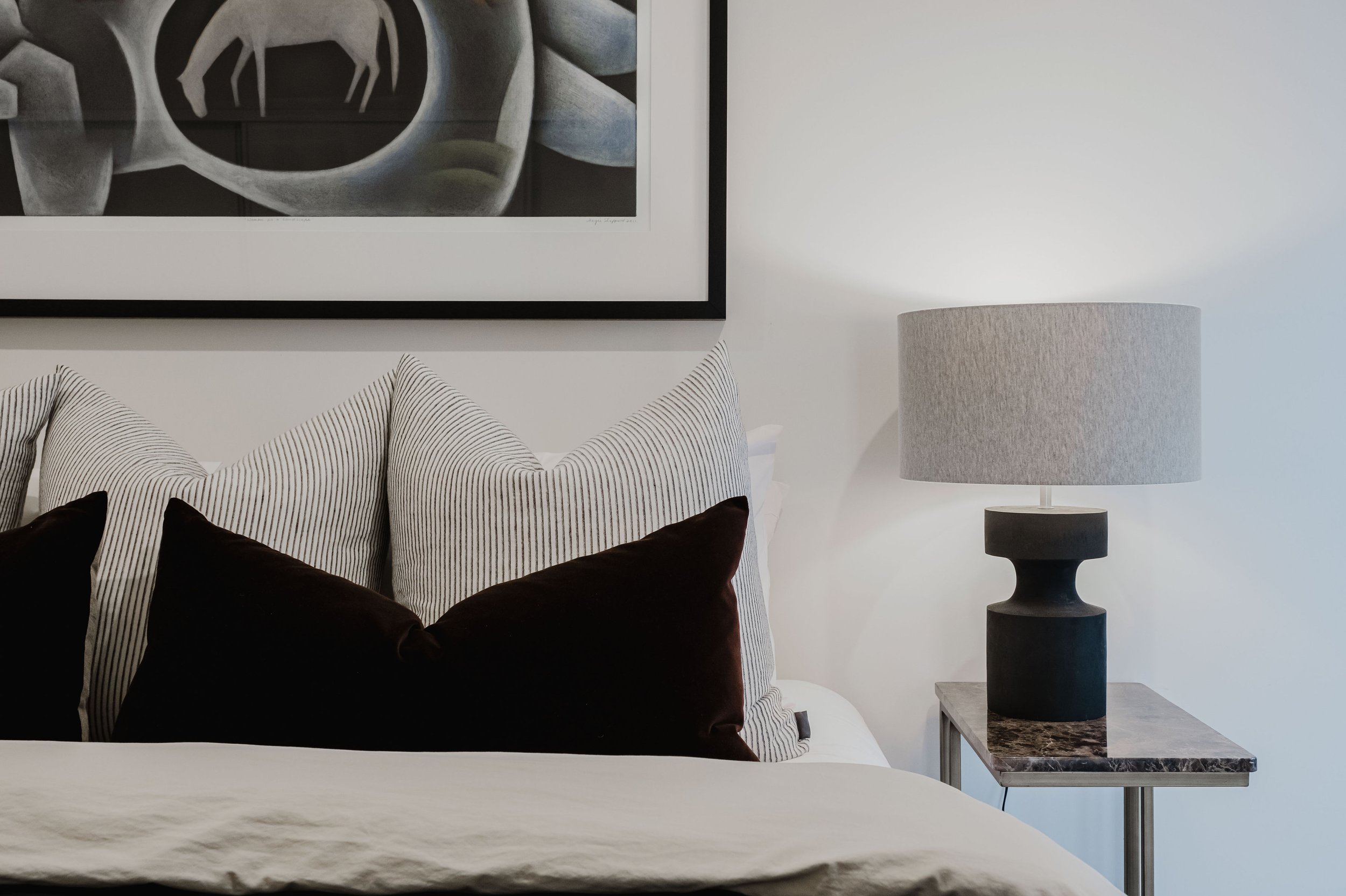 Close-up of a neatly made bed with decorative pillows, a modern table lamp on a bedside table, and framed artwork hanging above the bed in a stylish bedroom.