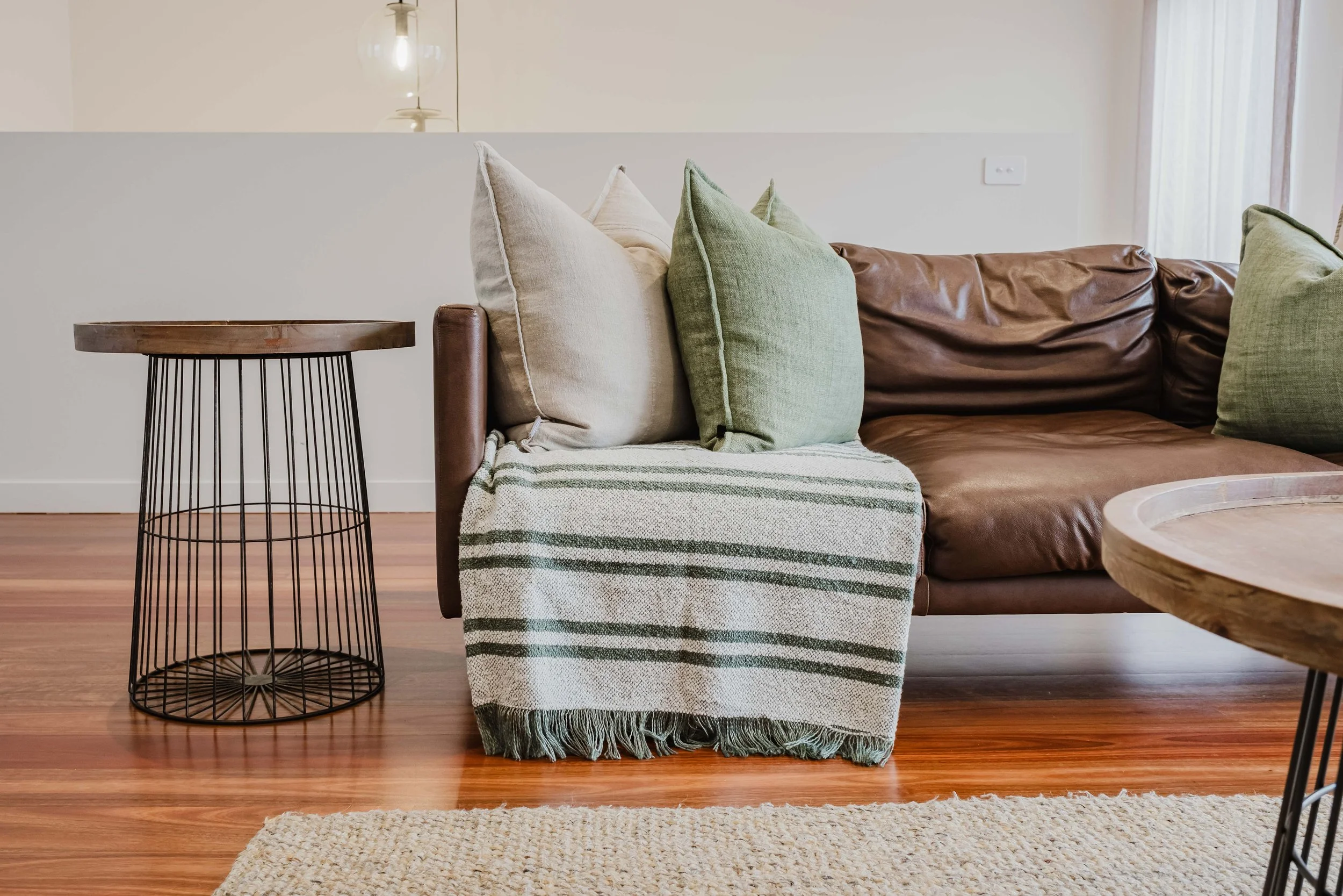Living room with a brown leather sofa with green and beige pillows, a striped throw blanket, a wooden side table with a wire base, a round wooden coffee table, hardwood floor, and a beige rug.