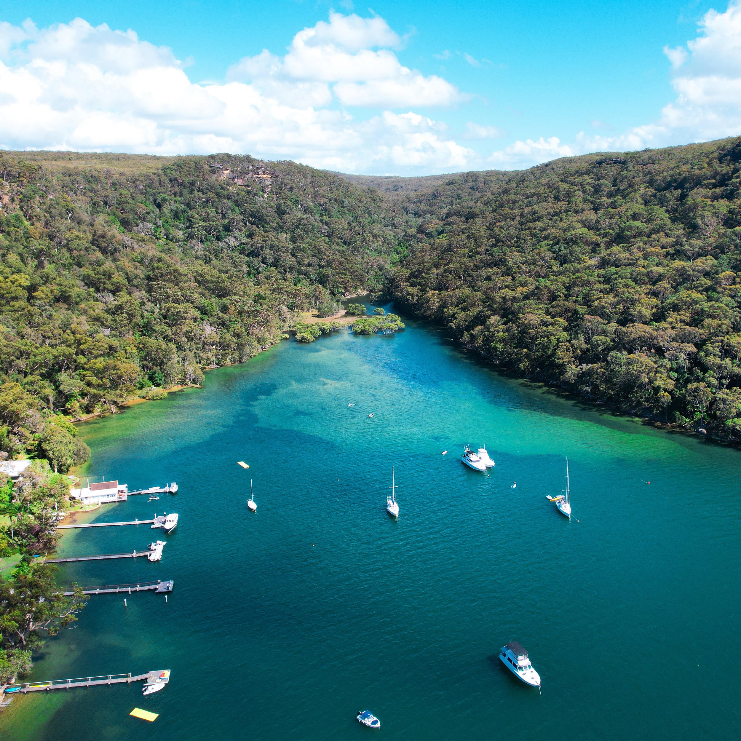 Aerial view of a Pittwater with boats, surrounded by lush green forests and clear blue skies.