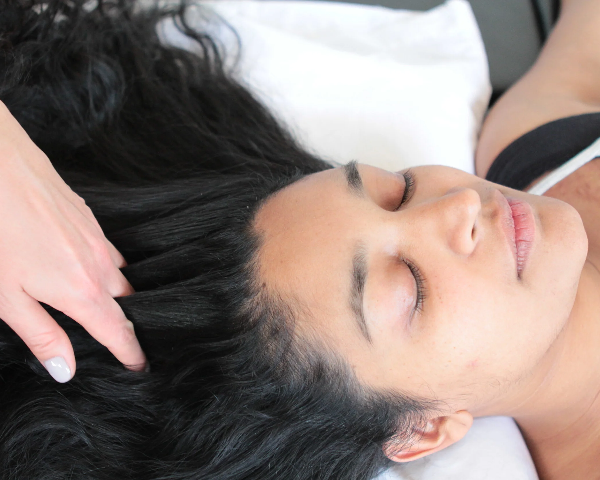 Close-up of a woman with long dark curly hair resting her head on a massage table with dark hair. The woman's eyes are closed and she appears peaceful.