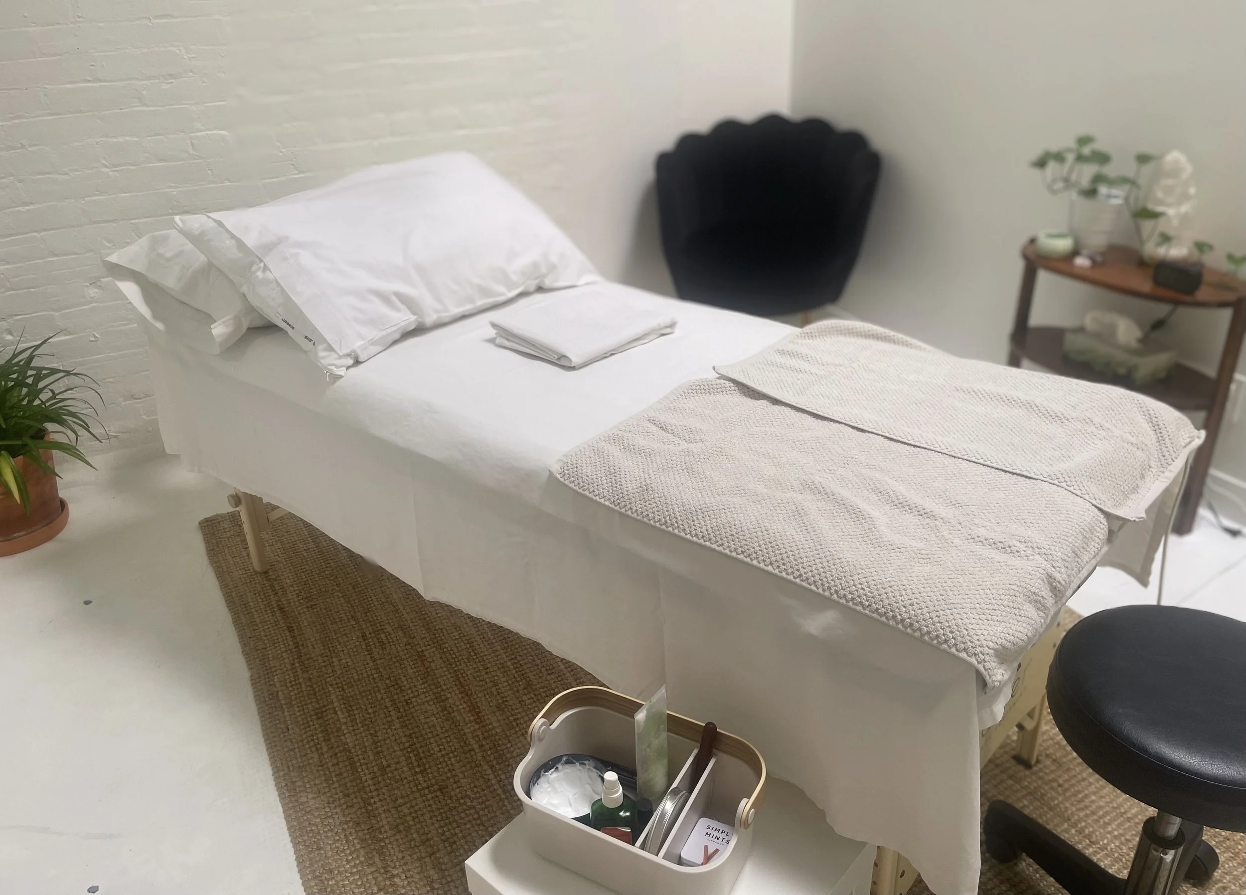 Massage or treatment table with white linens and a folded towel, set in a room with plants and a small side table holding wellness items.