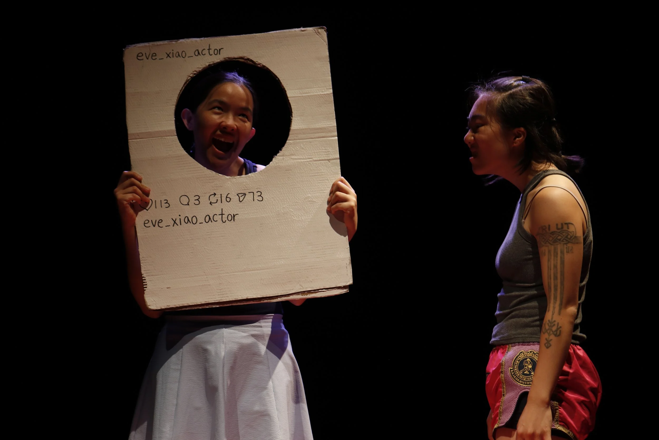 JJ Lam, the hatee, holds up a cardboard social media prop while Gawa Leung, the hater, scowls