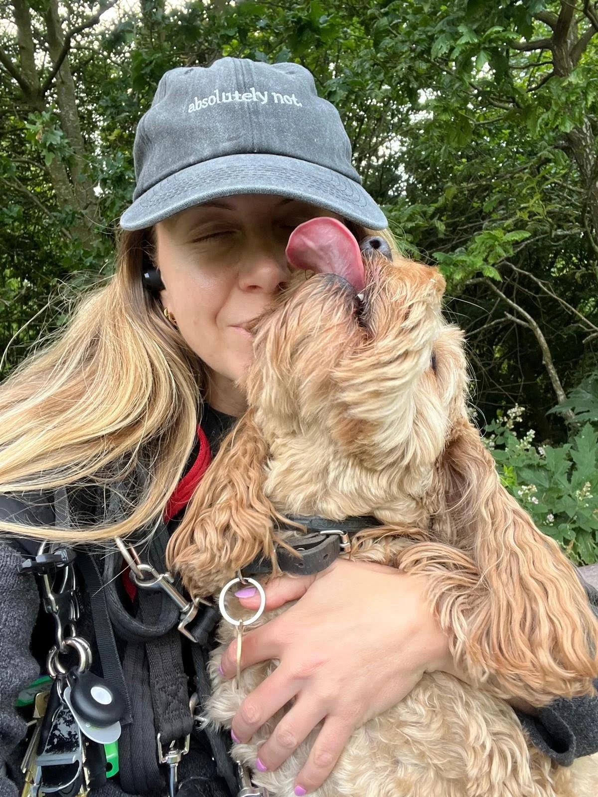 A woman wearing a gray cap with the words 'absolutely not' on it, hugging and kissing a golden cocker spaniel with its tongue out. They are outdoors with green foliage in the background.