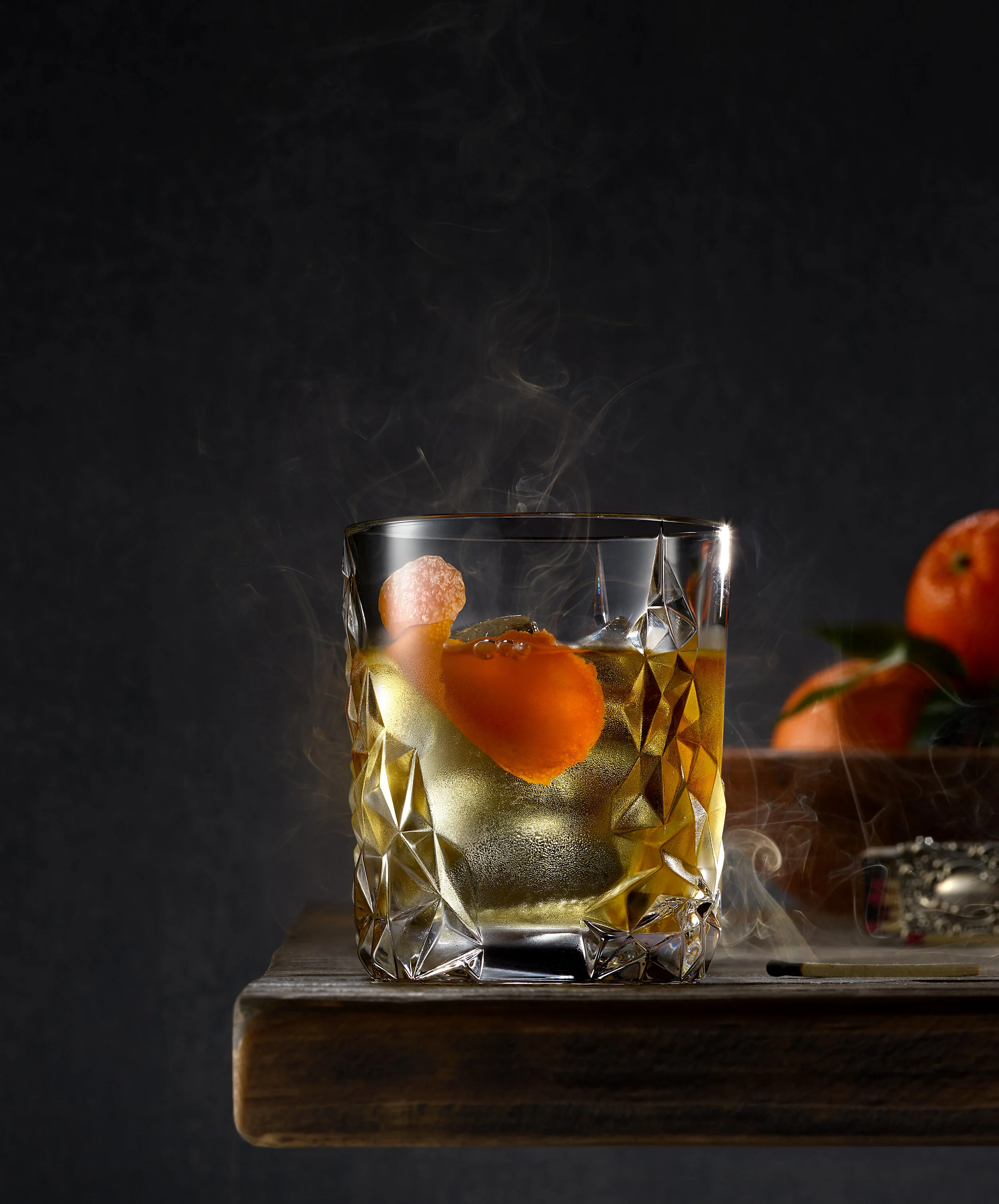 Glass with cocktail and orange peel on wooden surface with oranges in background.