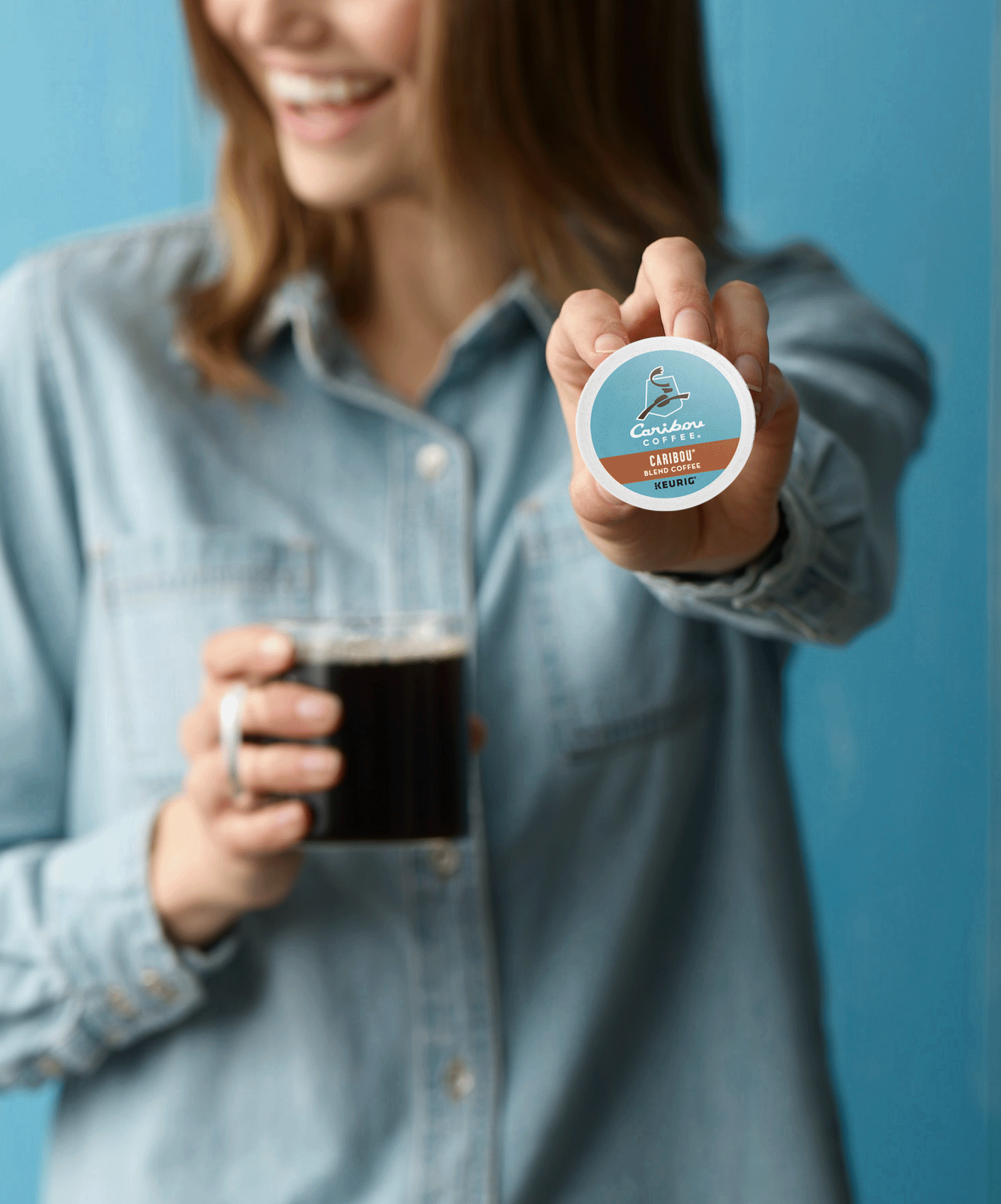 Person showing Caribou Coffee K-Cup and holding a mug of coffee, wearing denim shirt, in a blue background.