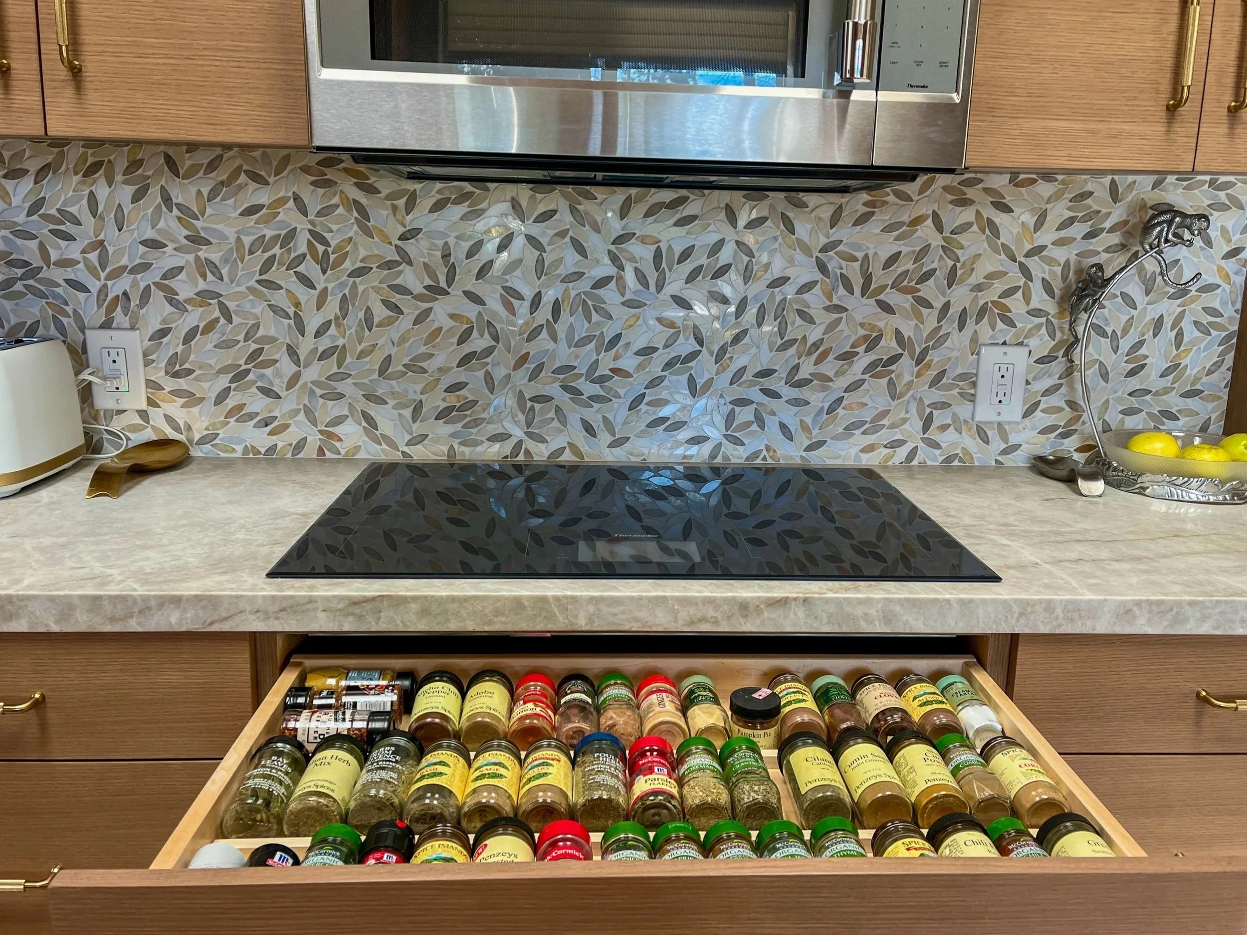Open kitchen drawer with various spices in small jars, countertop with a stove, backsplash with patterned leaves, microwave, and kitchen essentials in a kitchen remodel by Scott & Tim Management Group.