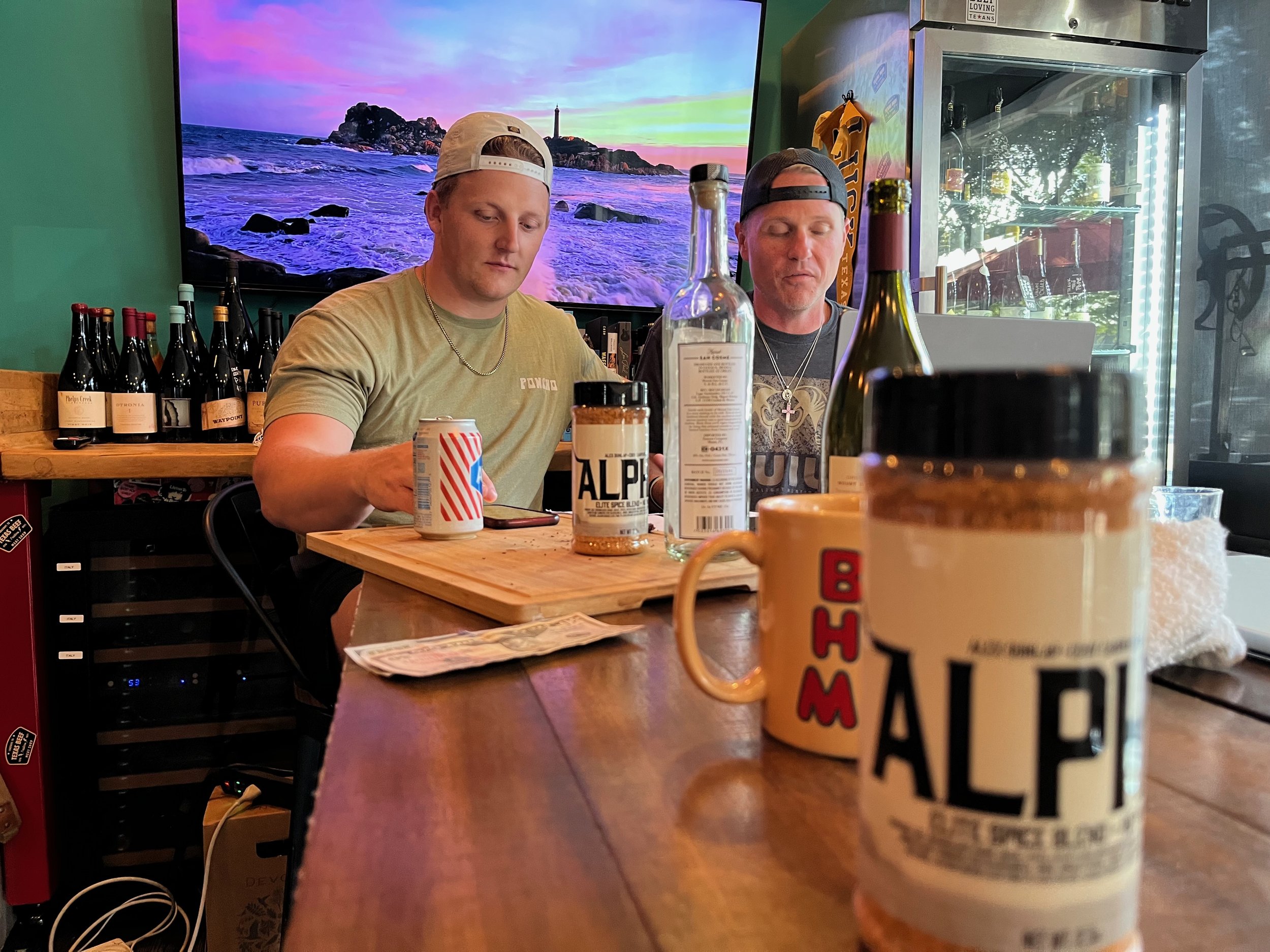 Two men sitting at a bar counter with a TV screen displaying a landscape of a lighthouse on a rocky island at sunset in the background. There are various bottles and cans on the counter, and a refrigerator with bottles inside.