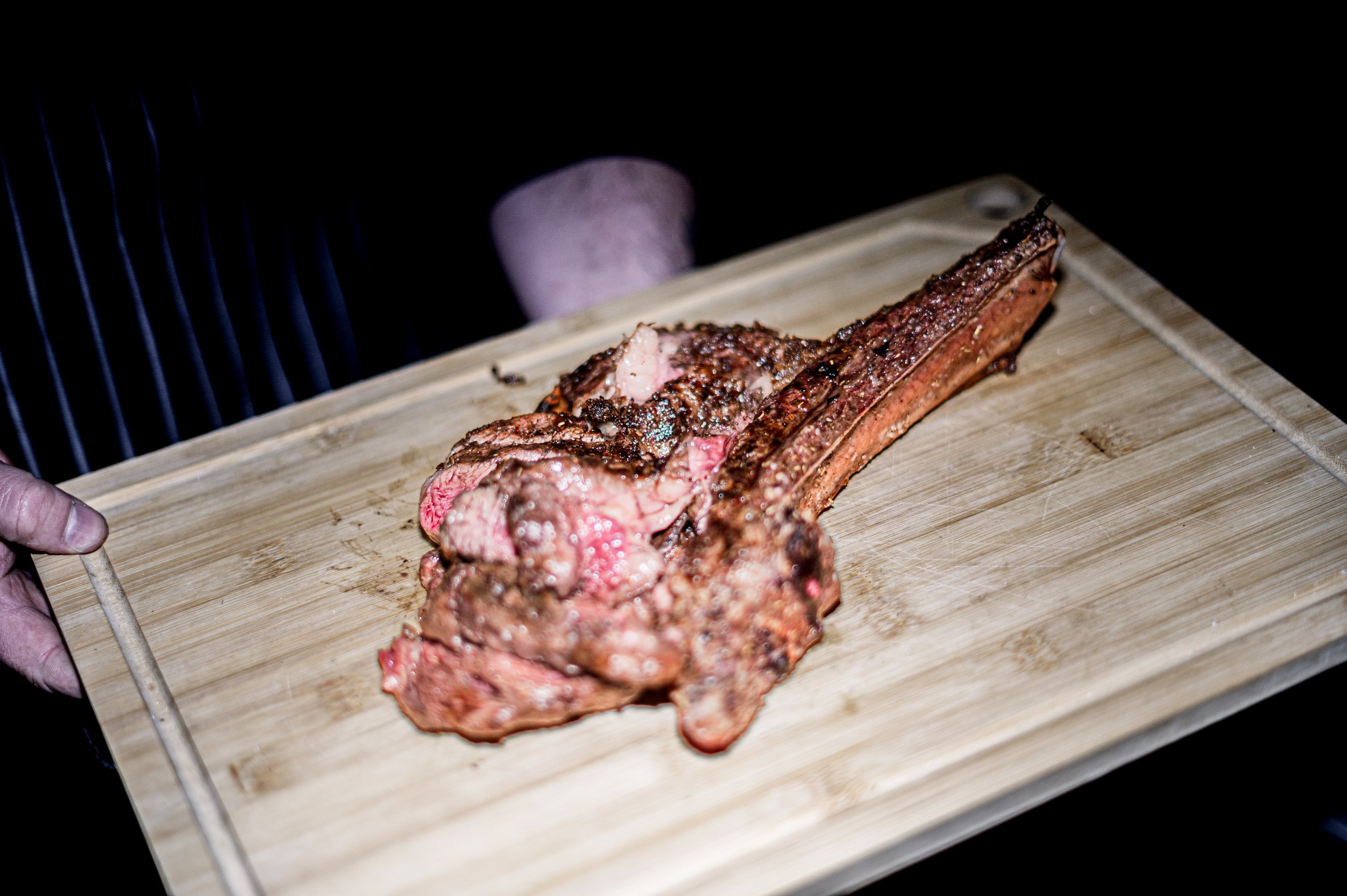 A person holding a wooden board with a cooked, seasoned ribeye steak on it.