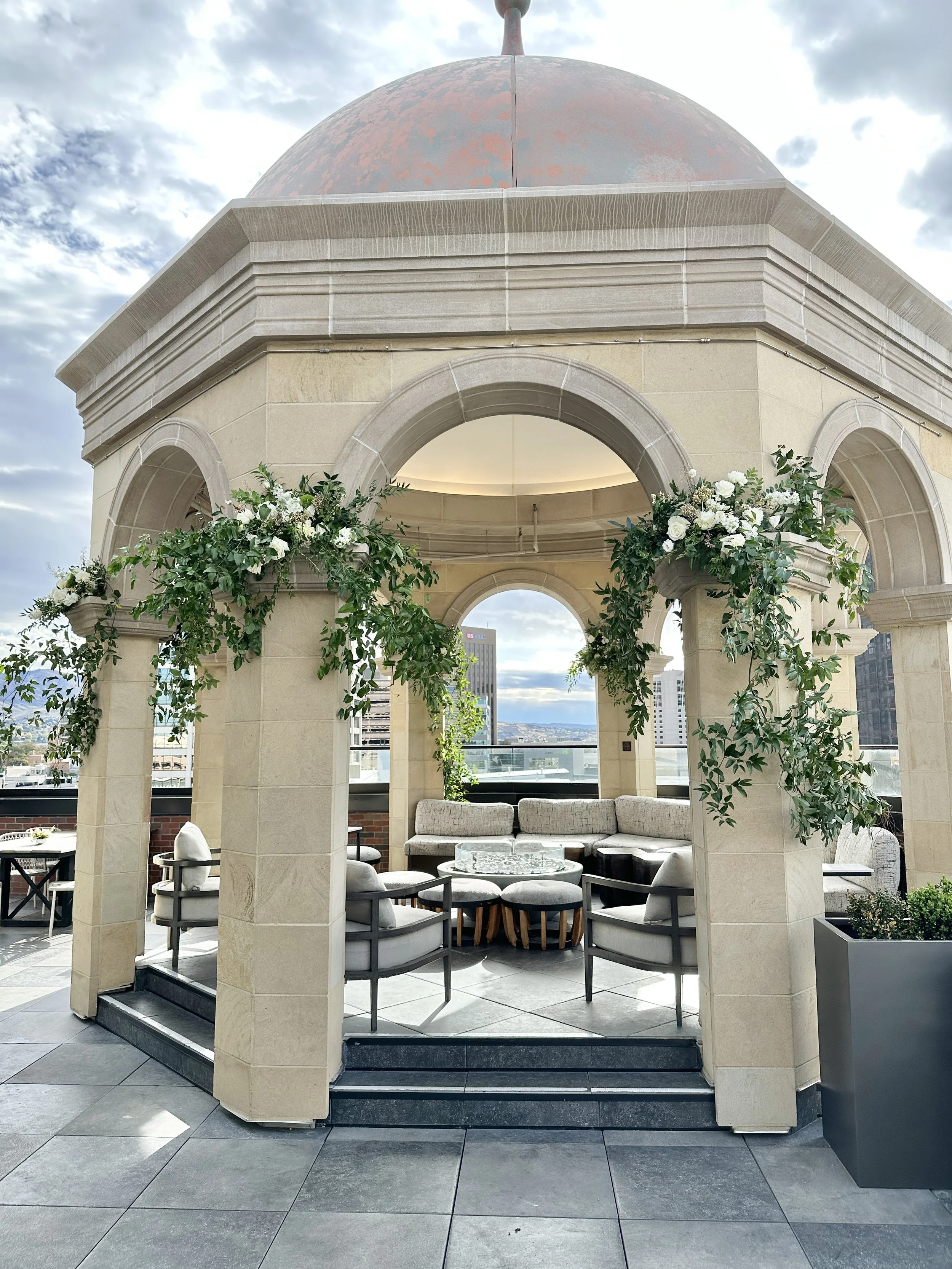 Outdoor rooftop wedding reception area decorated with white flowers and green foliage, created by Boise wedding florist at Edwards Flowershop, surrounded by city buildings and a cloudy sky.