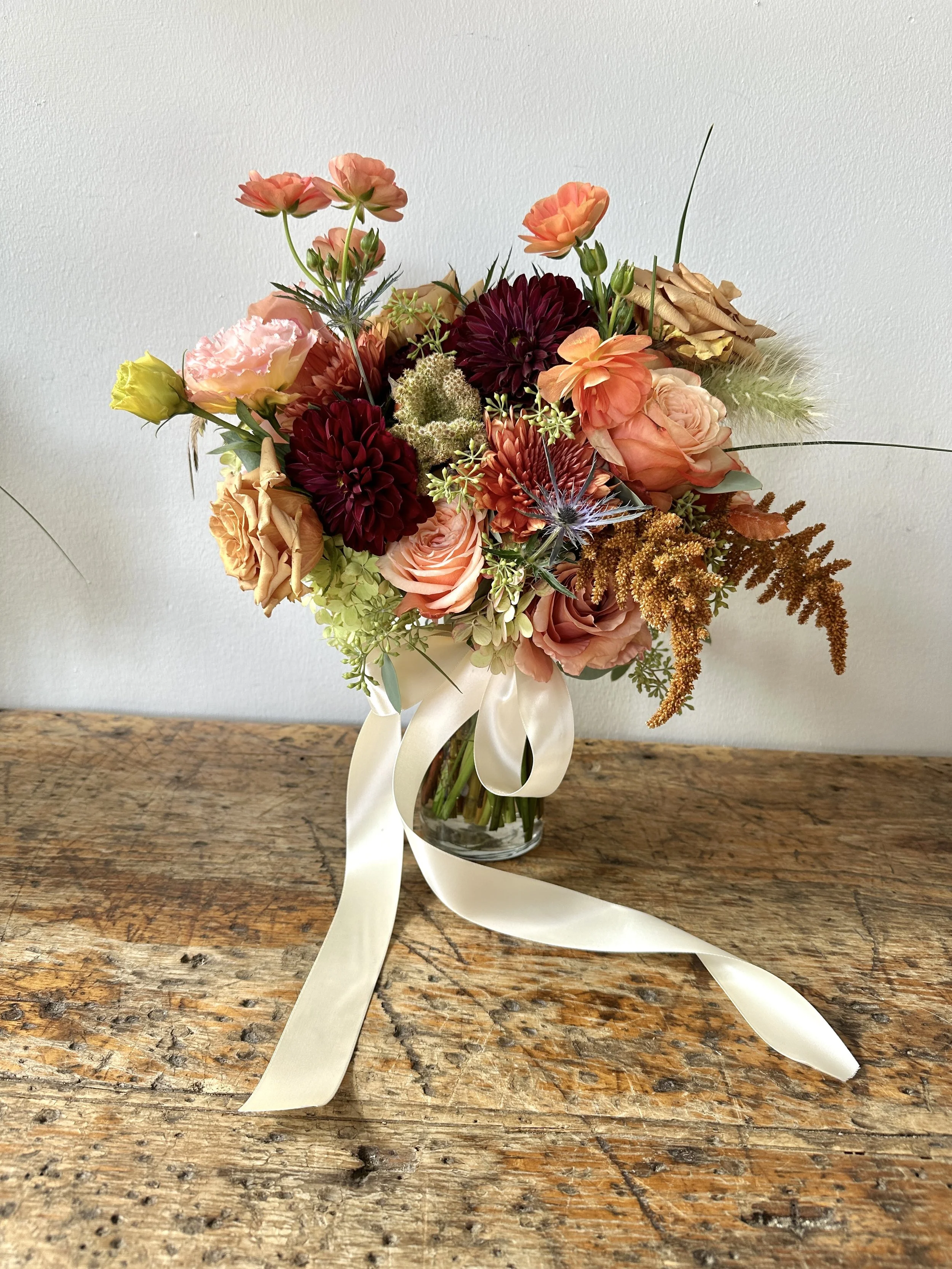 Boise wedding florist created bridal bouquet of mixed flowers in a clear glass vase with a cream-colored ribbon, placed on a rustic wooden table against a plain white wall. 