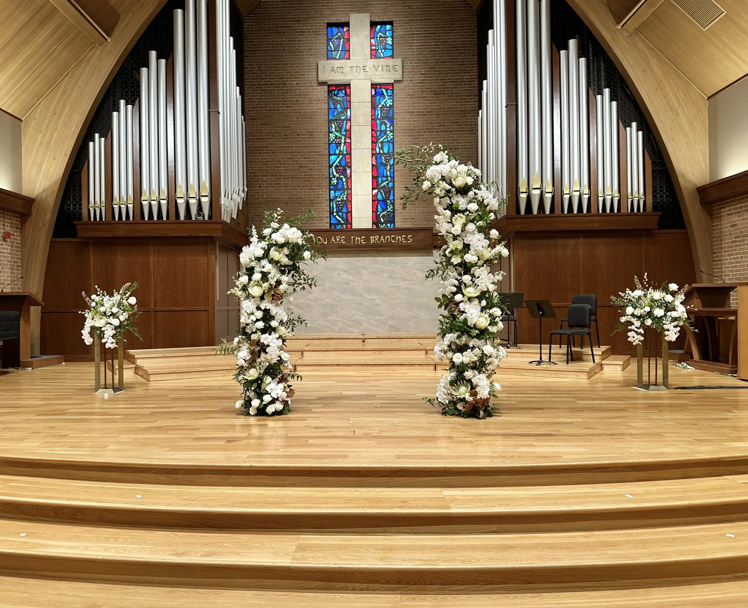 A large arch backdrop for a church wedding ceremony, featuring white flowers and lush greenery, accented by two large matching floral arrangements on stands. Wedding flowers by Boise florist Edwards Flowershop.
