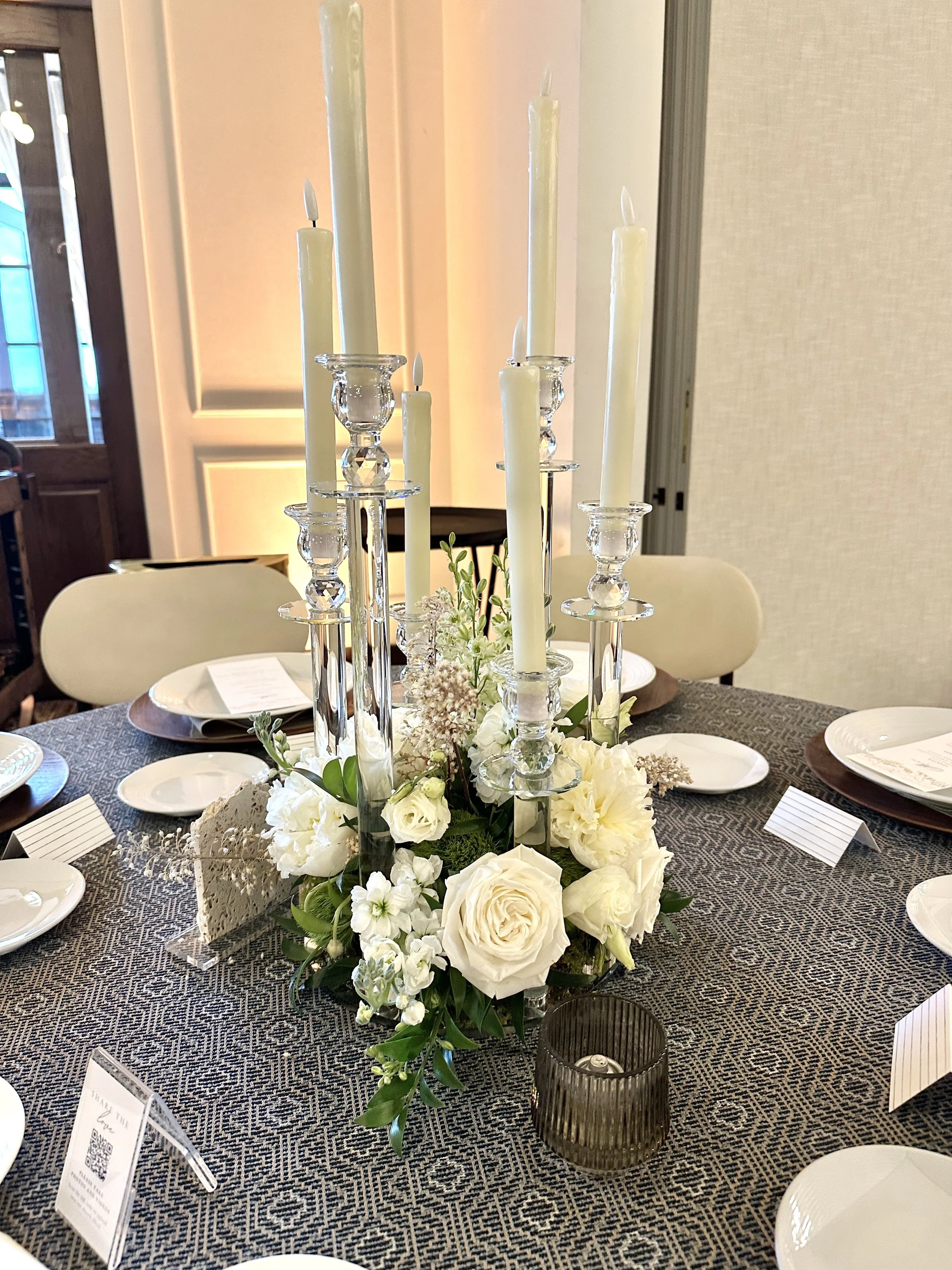 Elegant wedding table centerpiece with white roses, lilies, and greenery arranged around tall glass candle holders with white candles, set on a dining table with place settings and cards. Created by Boise wedding florist at Edwards Flowershop.