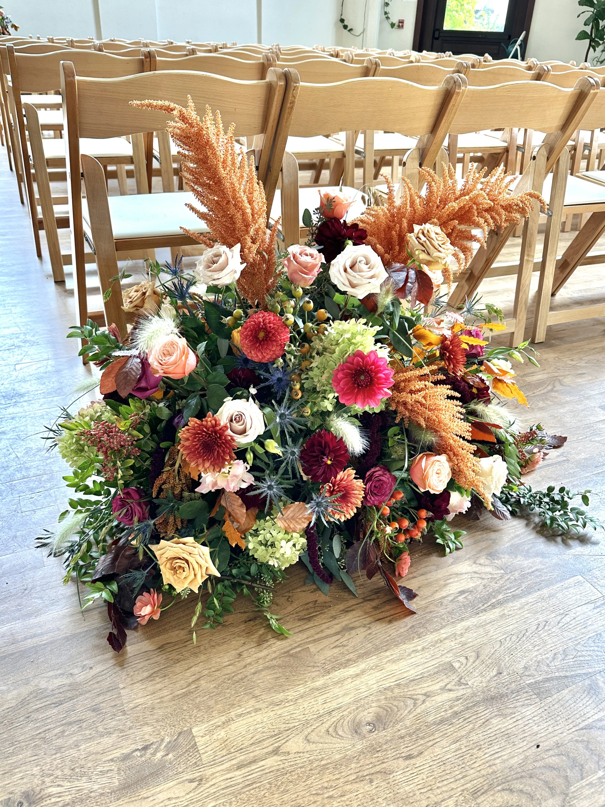 Wedding large arrangement of colorful flowers, including roses, dahlias, and greenery, sits in front of empty wooden chairs arranged in rows inside a well-lit room with wooden flooring and large windows. Boise wedding florist Edwards Flowershop.