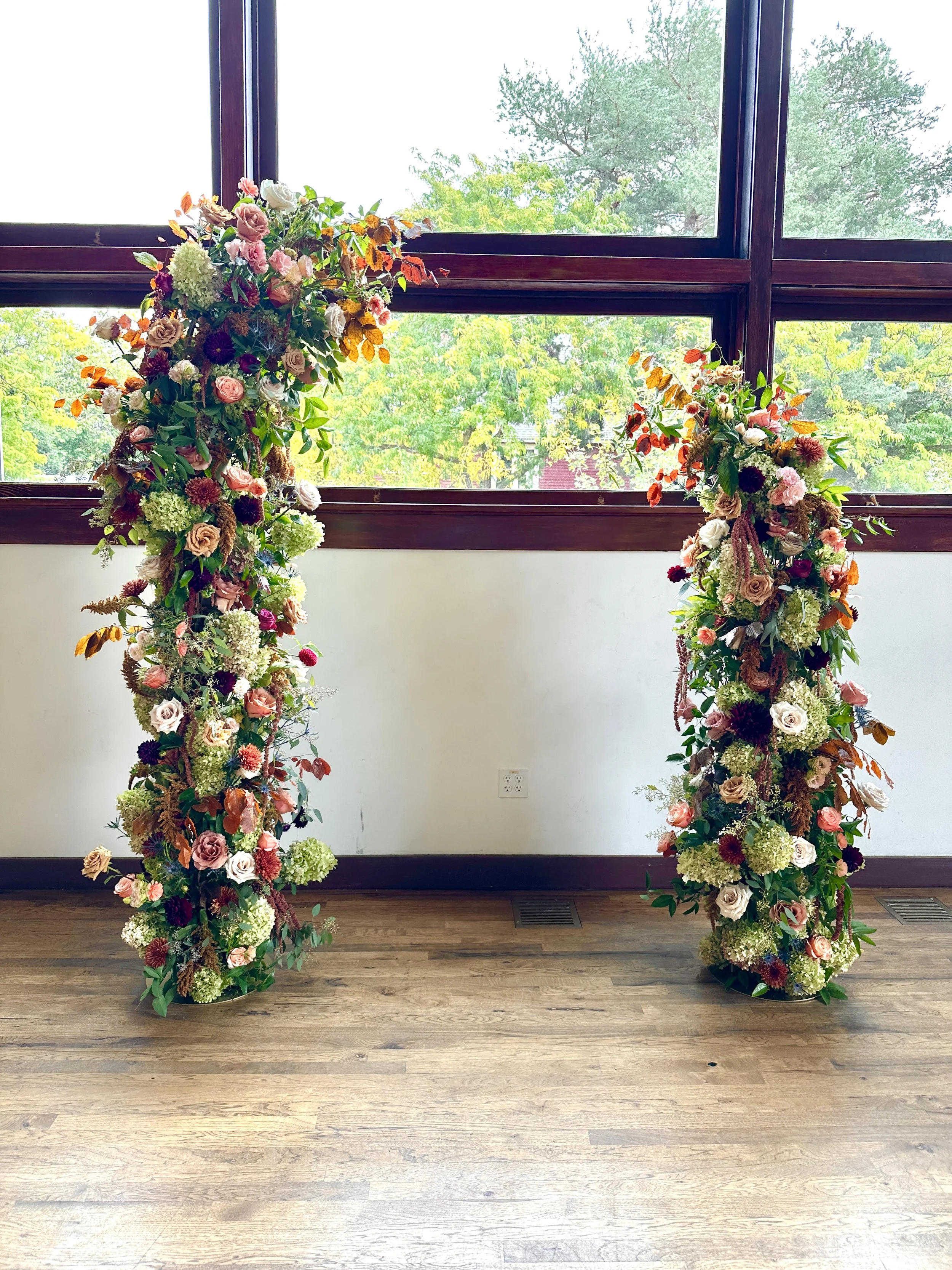 A two piece floral wedding backdrop with pink, white, purple, and green flowers and leaves, set on the floor in front of large window with trees outside, indoors on a wooden floor. Created by Boise wedding florist at Edwards Flowershop.