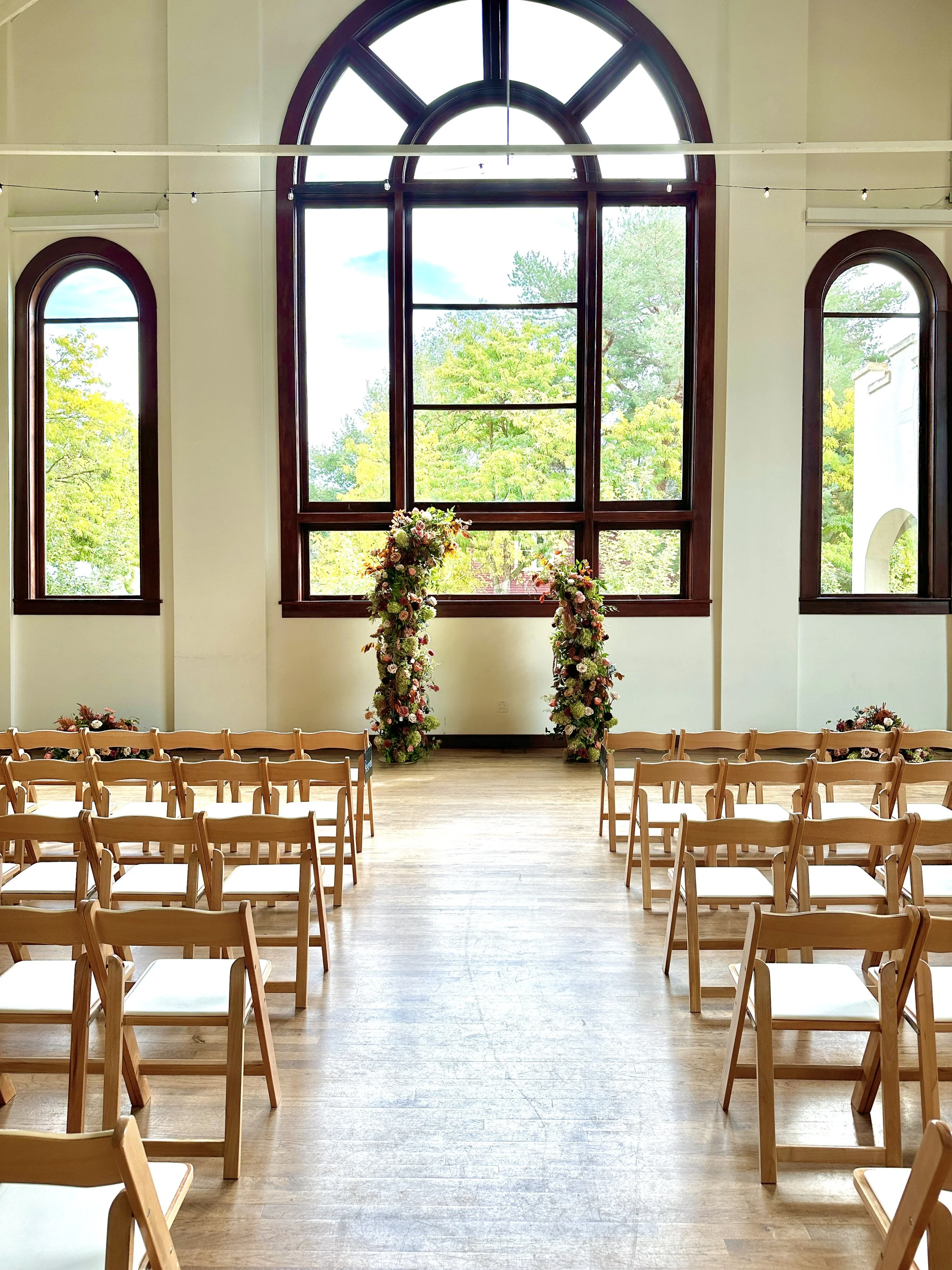 A two piece floral wedding backdrop with pink, white, purple flowers in a ceremony space with wooden chairs arranged in rows and floral decorations near the large window. The space has high ceilings, large arched windows, and lots of natural light.