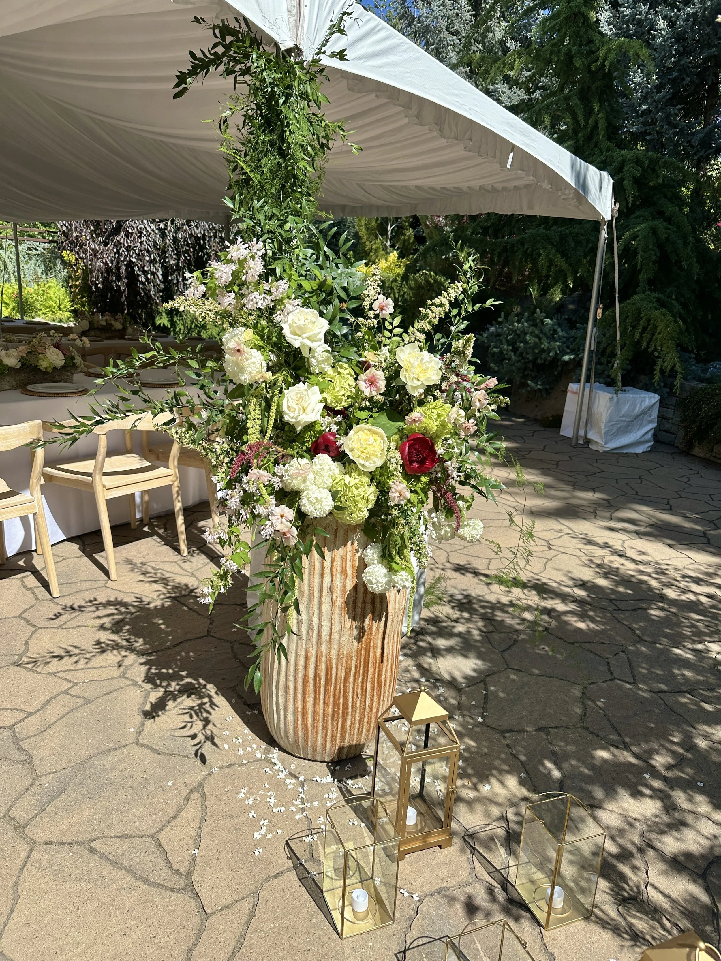 Wedding reception flowers by Boise florist Edwards Flowershop featuring a tall floral arrangement with white, pink, and red flowers and green foliage in a beige, textured vase under a tan canopy, with lanterns placed on the ground nearby. 