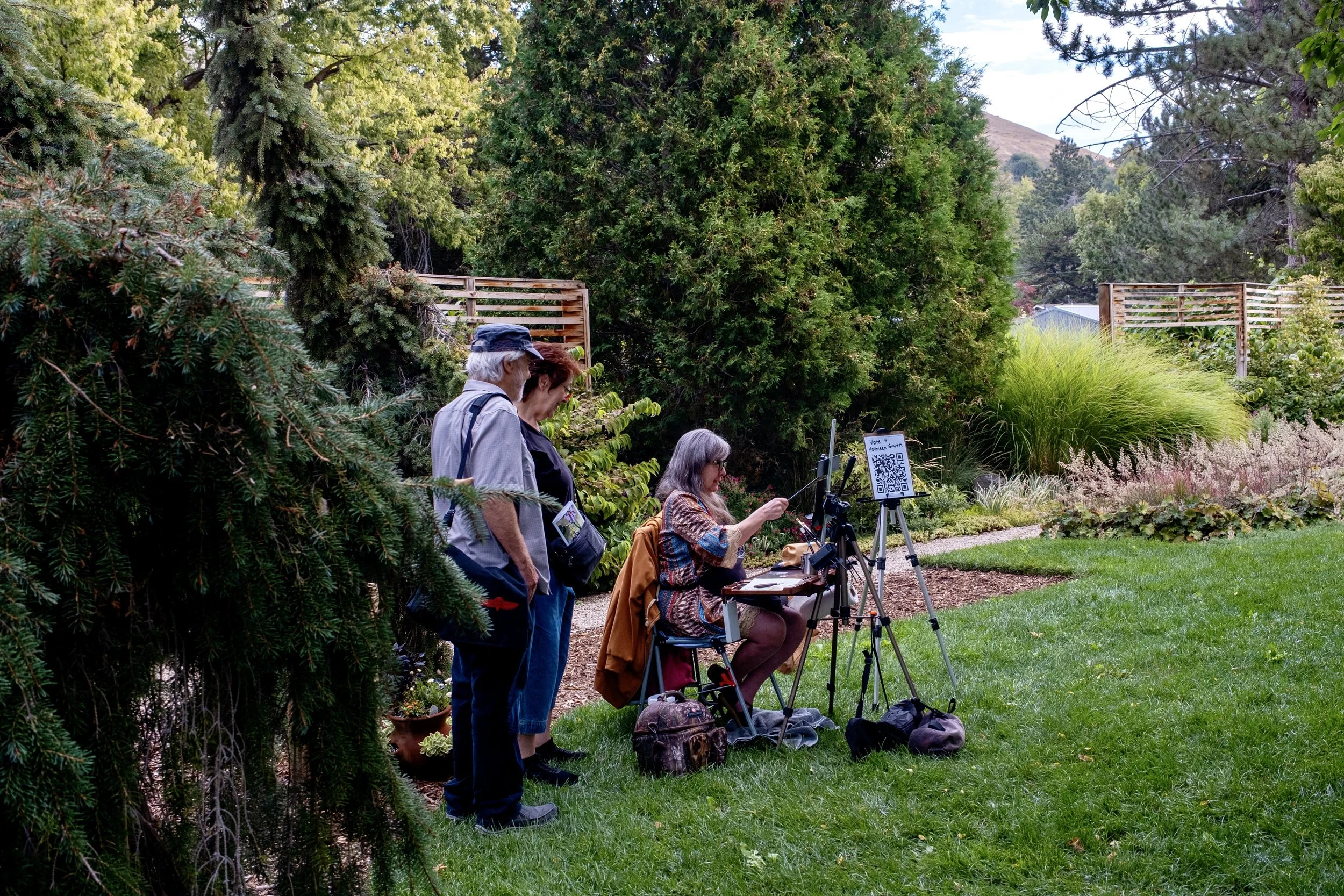A woman sits pointing in a lush green garden. Two people stand beside her, observing.